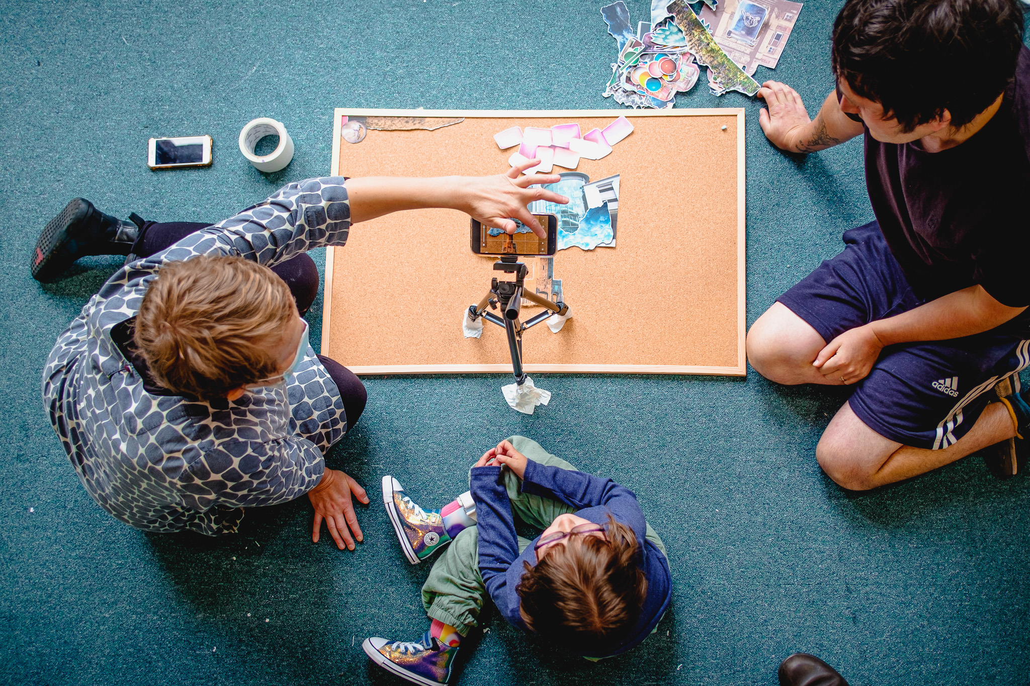 Three people sit on the floor around a pinboard with paper, art supplies, and a mobile mounted on a tripod, appearing to work on a creative project together.