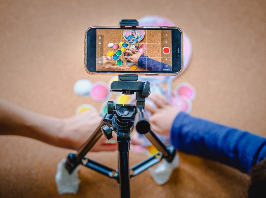 A smartphone on a tripod records two people’s hands playing a colourful board game, with tokens and pieces spread on a brown surface.