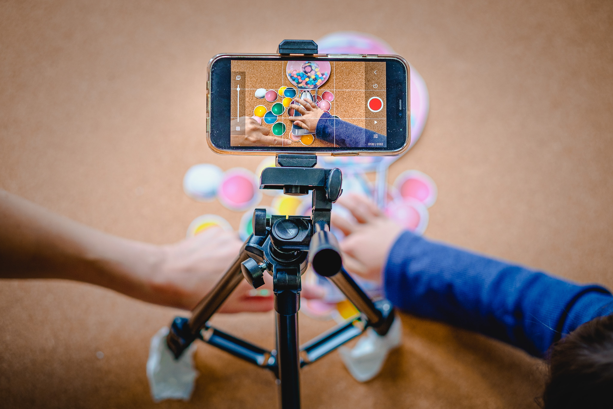A smartphone on a tripod records two people’s hands playing a colourful board game, with tokens and pieces spread on a brown surface.