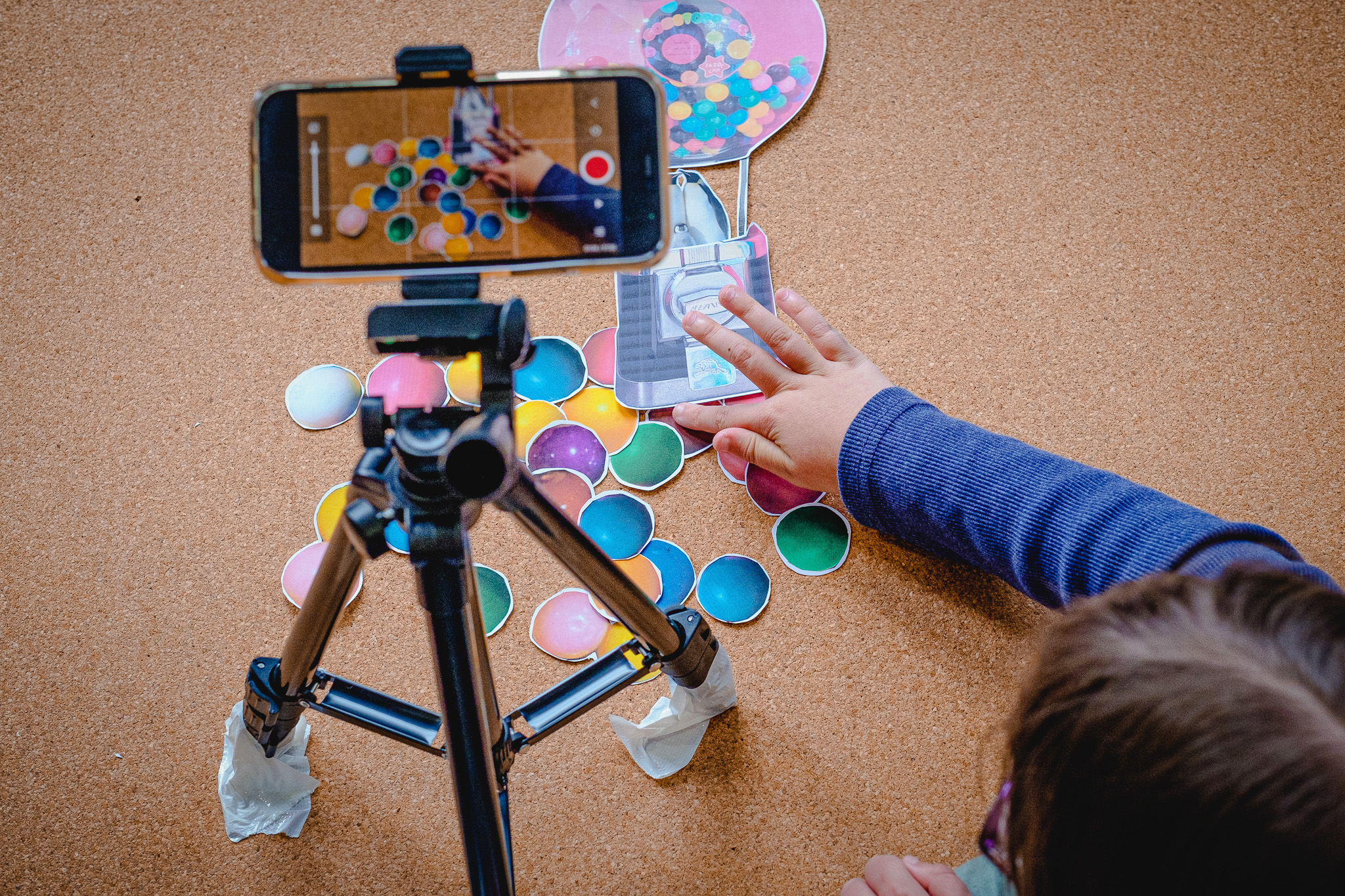 Child's arm arranging colourful paper circles on corkboard with a smartphone on a tripod recording the activity.