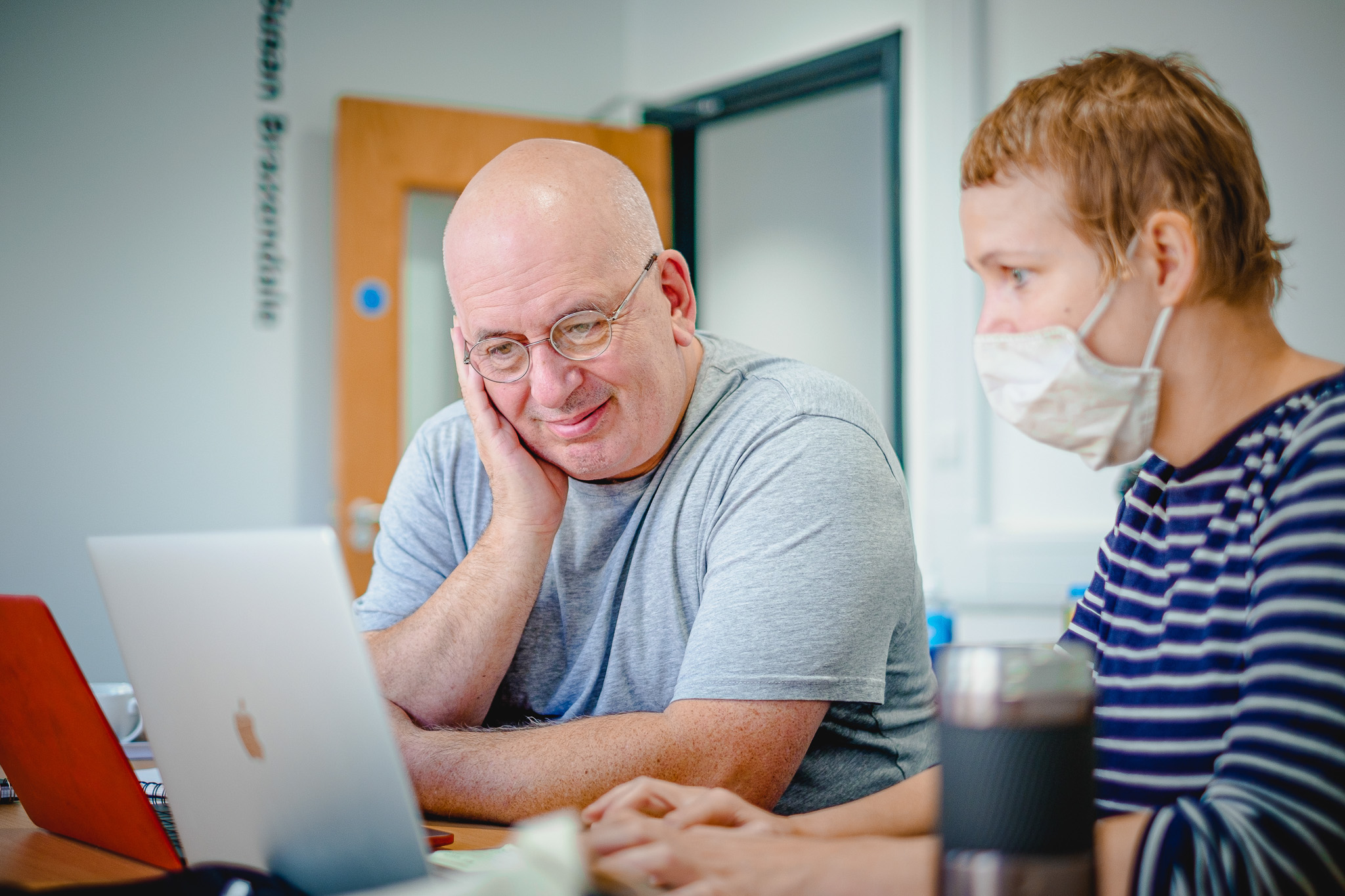 Two people work together at a table with a laptop. One person, wearing glasses and a grey shirt, smiles at the screen. The other, in a striped shirt and face mask, is focused on the laptop.