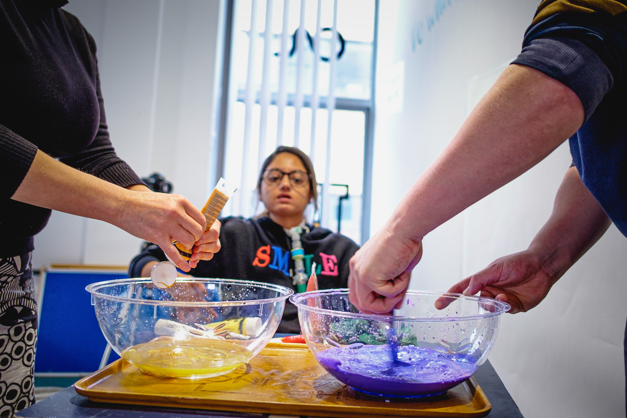 Two people mix ingredients in separate bowls containing yellow and purple liquids on a table. A person in a black 