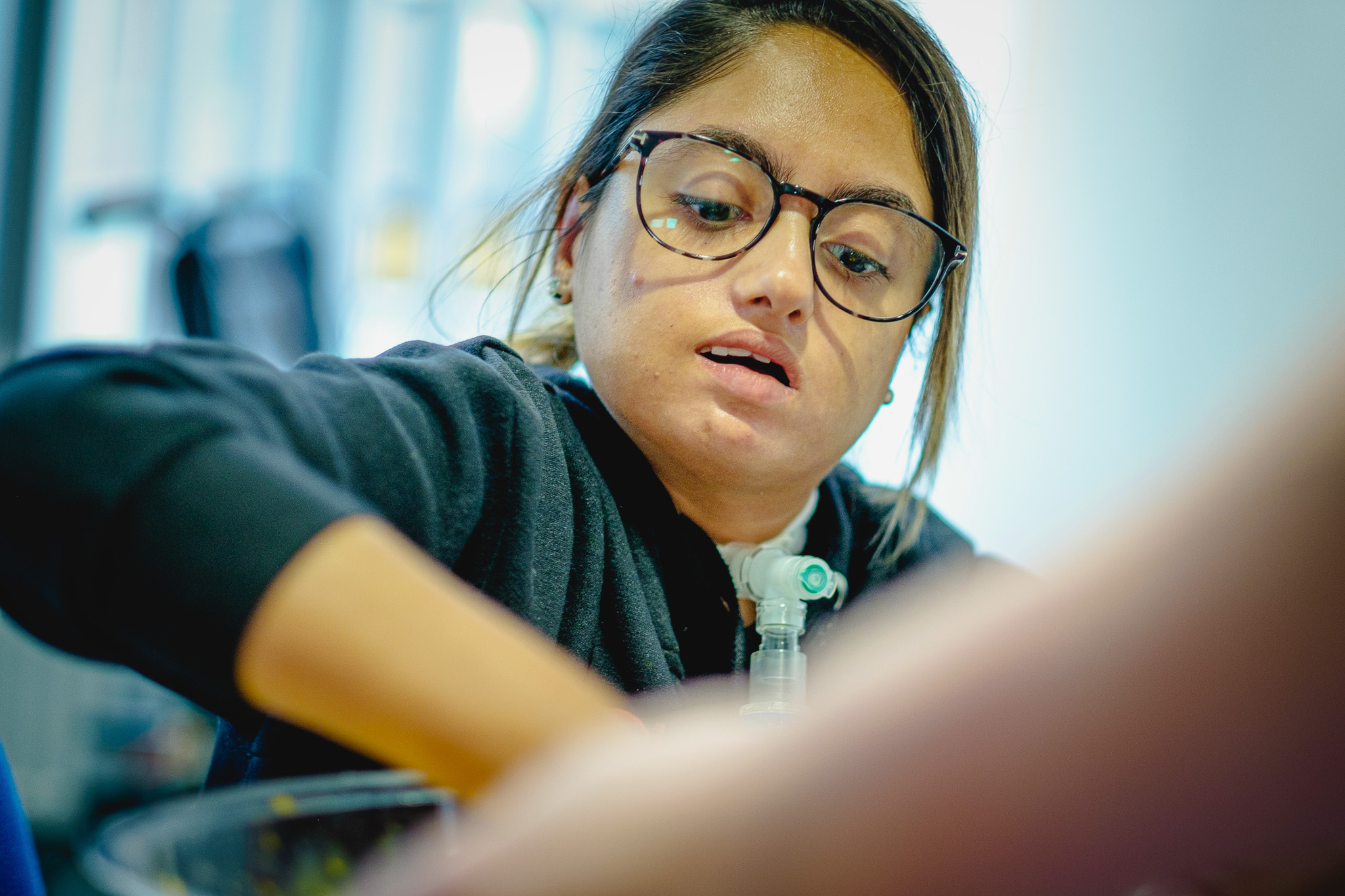 A woman with glasses reaching forward. She's wearing a black top and a medical device is visible around her neck.