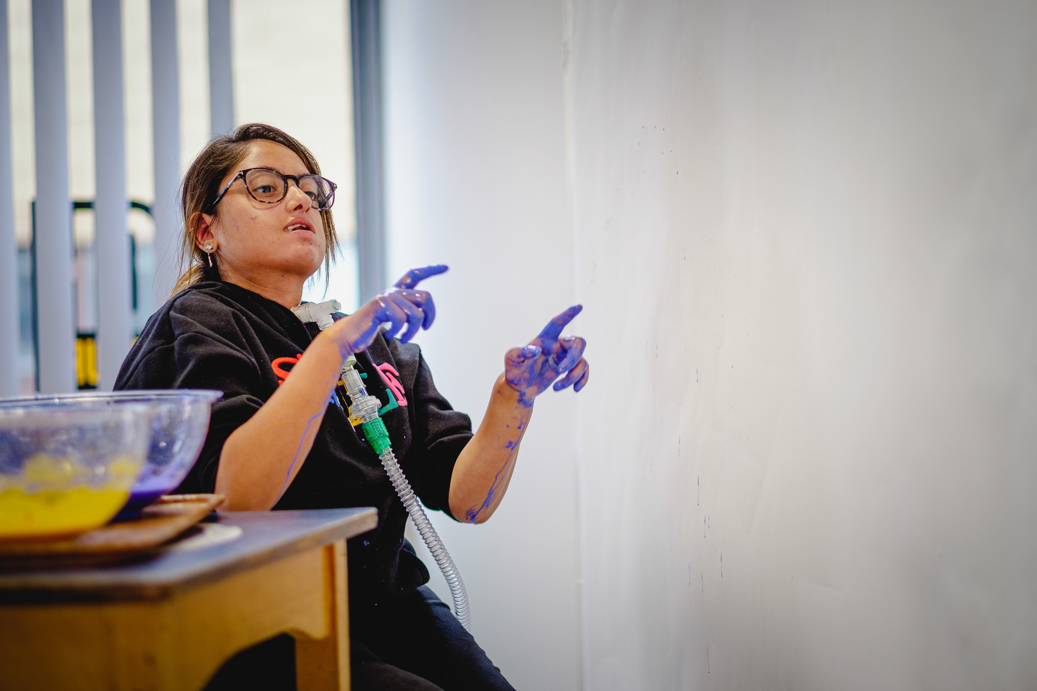 A person with glasses and a ventilator tube sits by a table, their fingers covered in blue paint, gesturing towards a blank white wall as if preparing to create artwork.