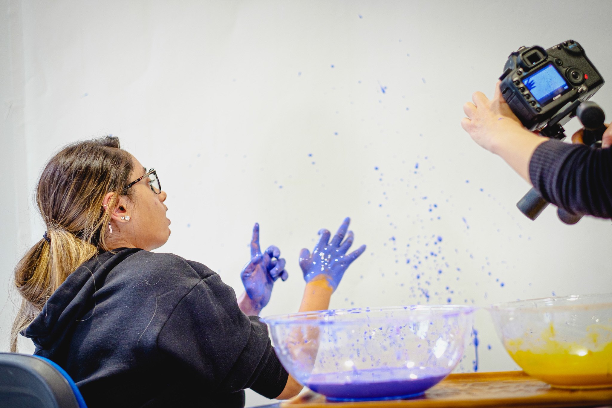 A woman with paint-splattered hands gestures toward the camera; blue paint splashes the wall, with two bowls of paint nearby.
