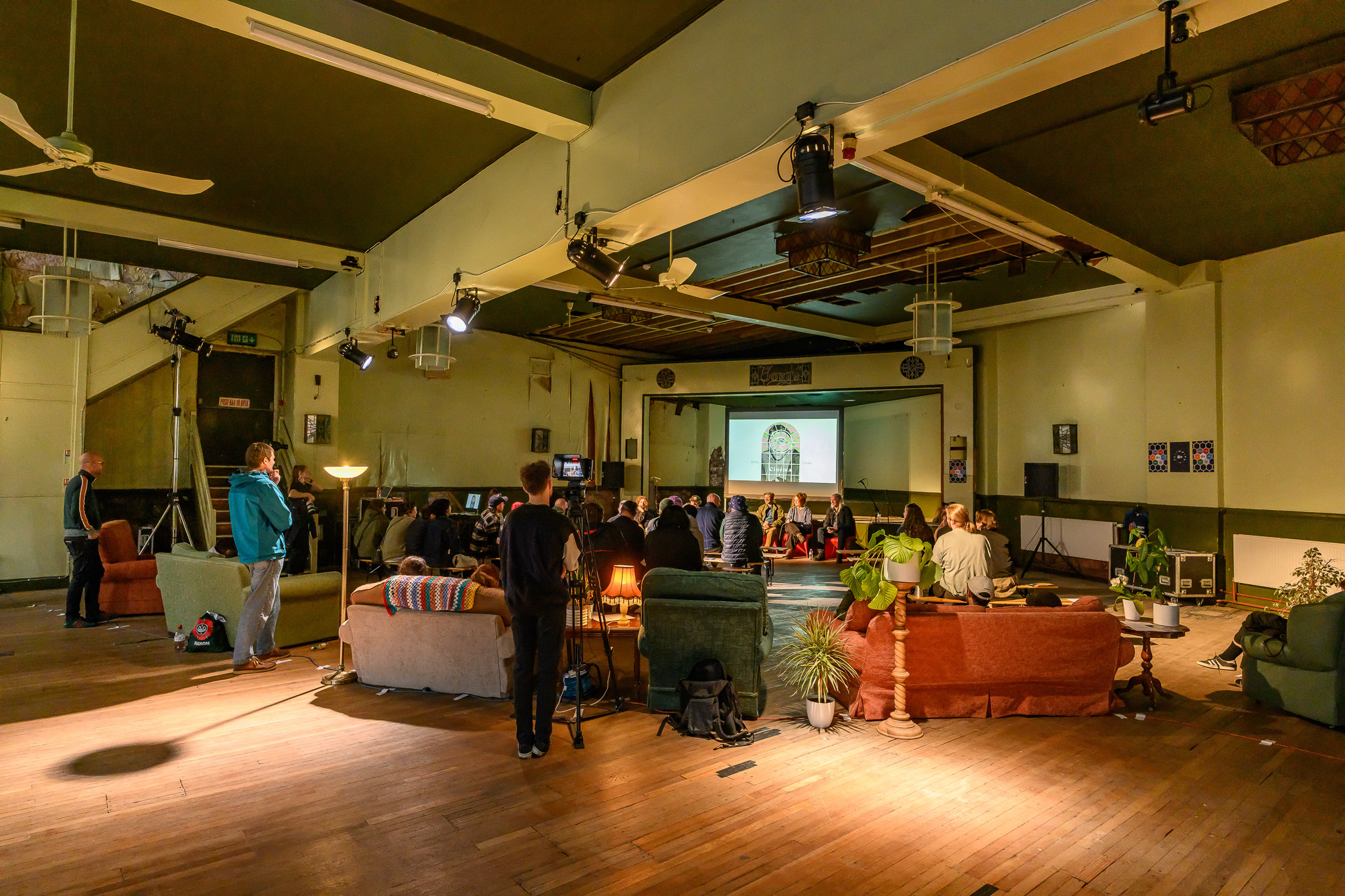 A group of people sits on sofas and chairs in a large hall, watching a panel talk. The room has warm lighting, wooden floors, and eclectic furniture.