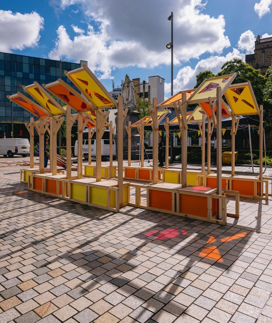 Colourful outdoor market stalls with yellow and orange panels and wooden frames stand on a tiled plaza. Shadows and sunlight create patterns on the ground; modern buildings and trees are visible in the background.
