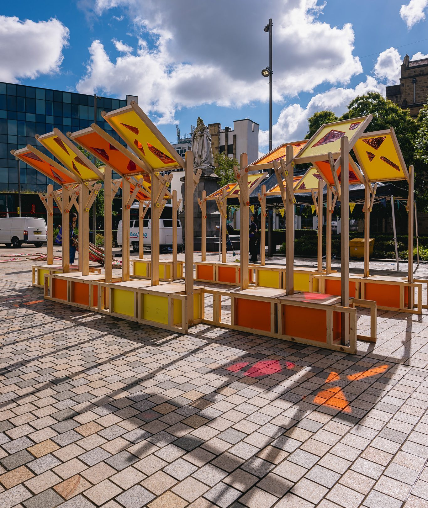 Colourful outdoor market stalls with yellow and orange panels and wooden frames stand on a tiled plaza. Shadows and sunlight create patterns on the ground; modern buildings and trees are visible in the background.