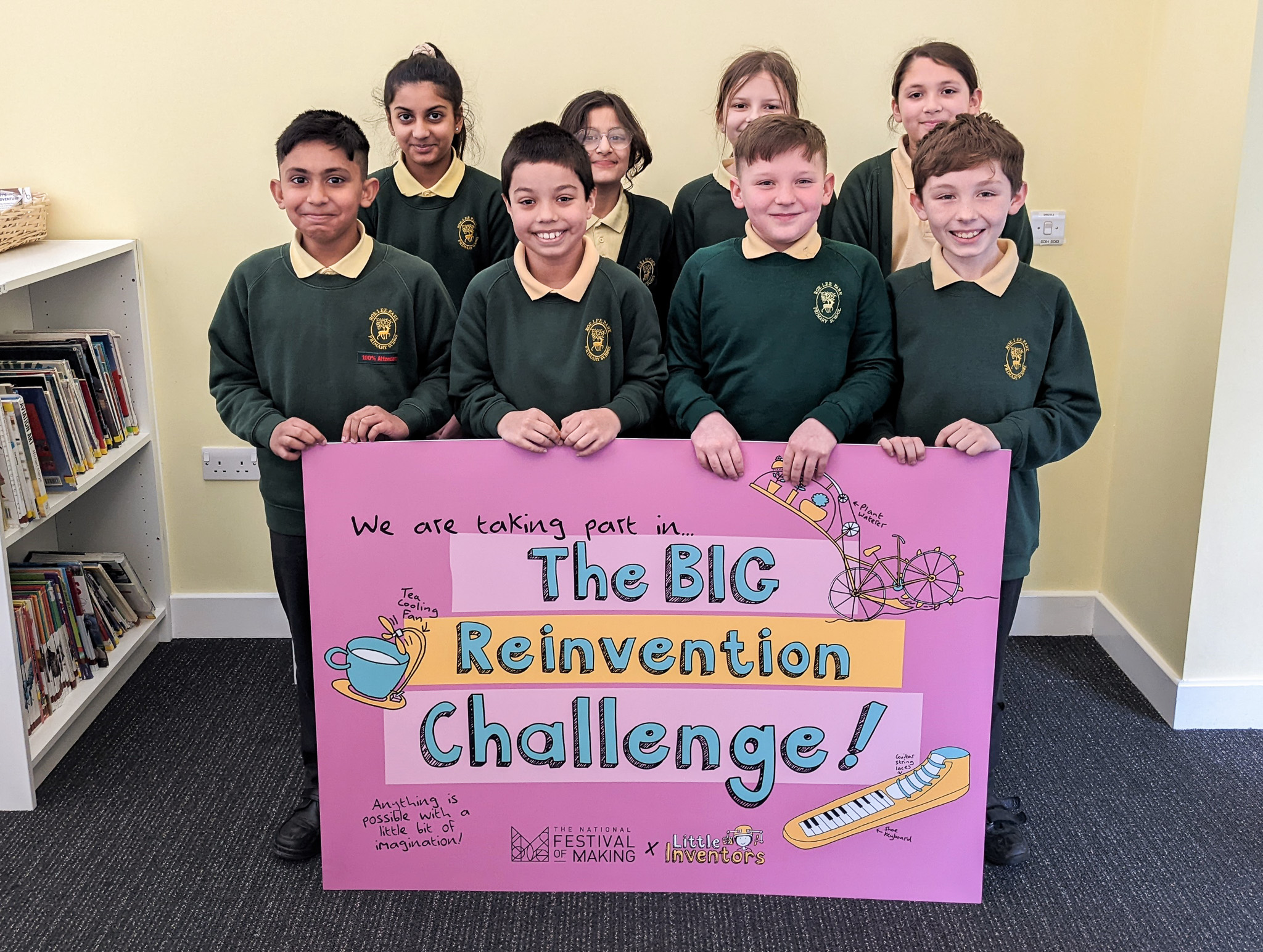 Seven children in green school uniforms stand indoors, smiling and holding a large pink sign that reads The BIG Reinvention Challenge! with drawings of inventions and the words, We are taking part in....