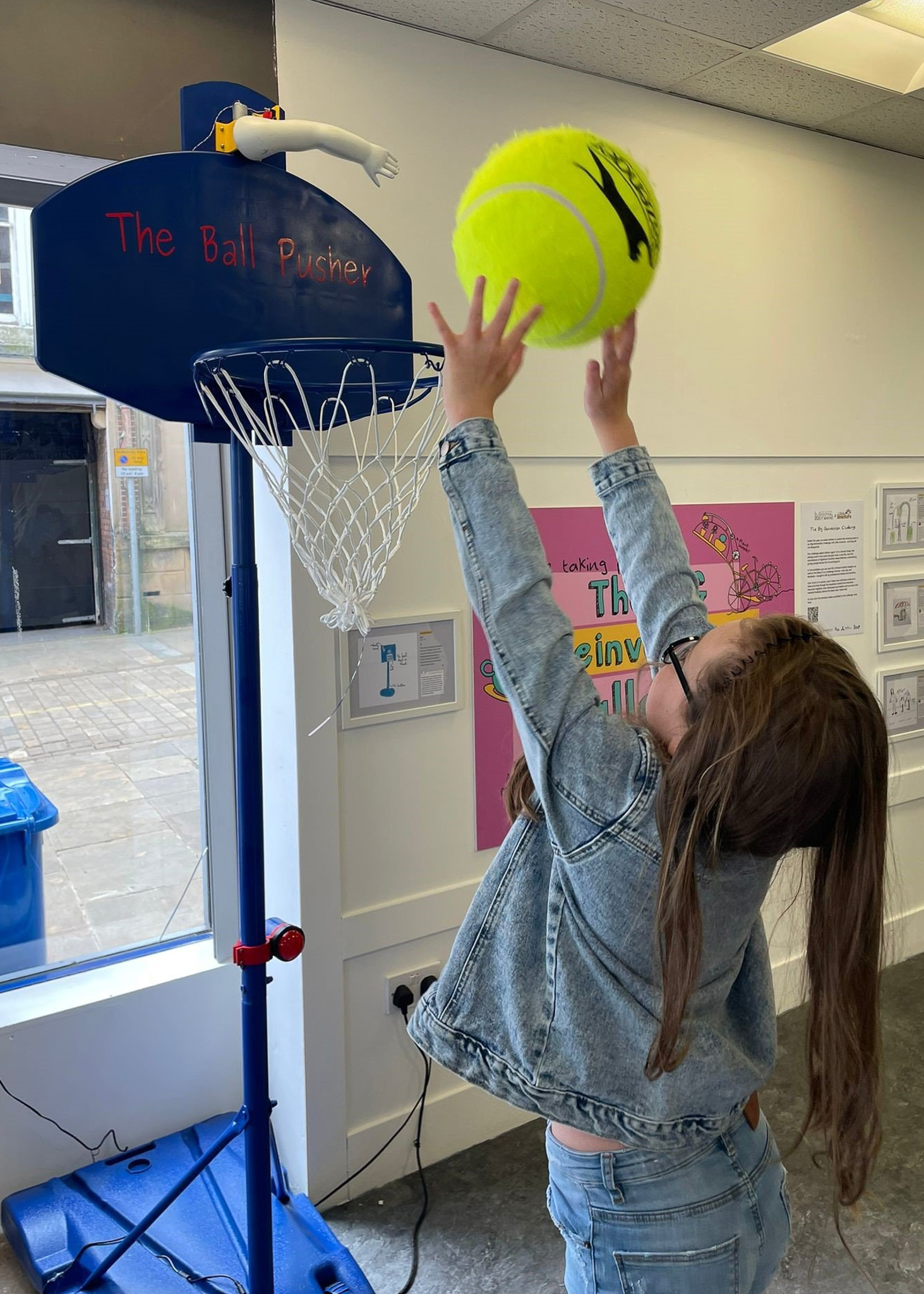 A young girl in a denim jacket reaches up with a large tennis ball, aiming for a basketball hoop labeled 