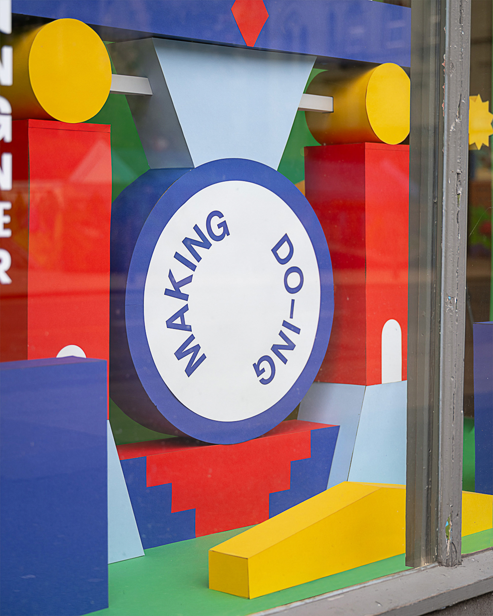 A colourful window display with geometric shapes and a circular sign in the centre that reads MAKING DO-ING in blue letters. The background includes red, yellow, and blue blocks and patterns.