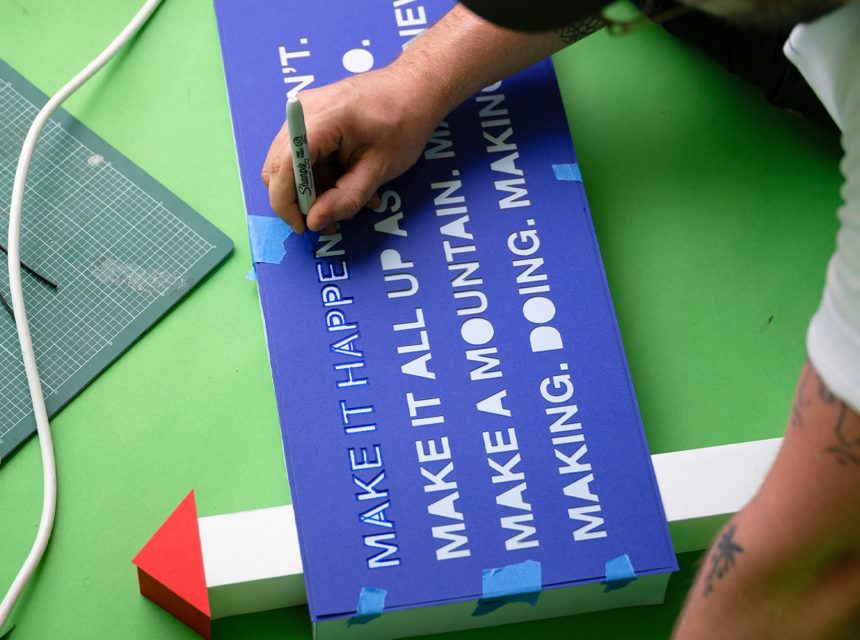 A person writes on a blue sign with white text using a black marker pen. The sign is taped to a white arrow with a red tip, and crafting tools and cables are nearby on a green surface.