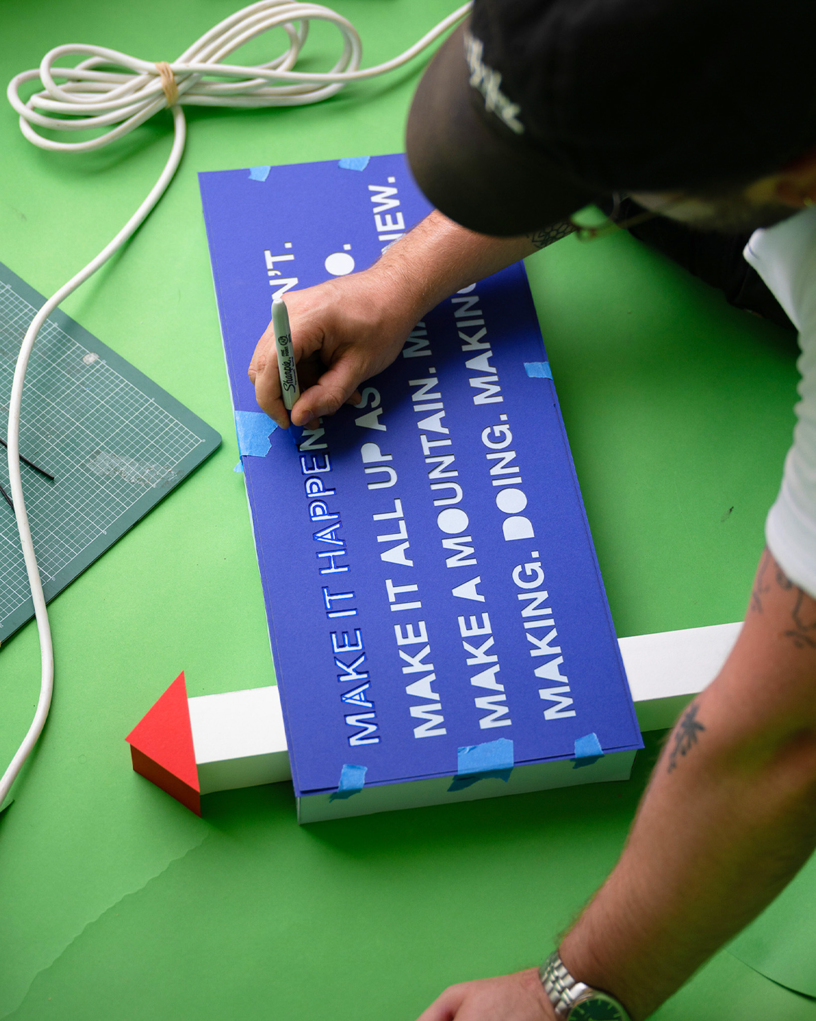 A person writes on a blue sign with white text using a black marker pen. The sign is taped to a white arrow with a red tip, and crafting tools and cables are nearby on a green surface.