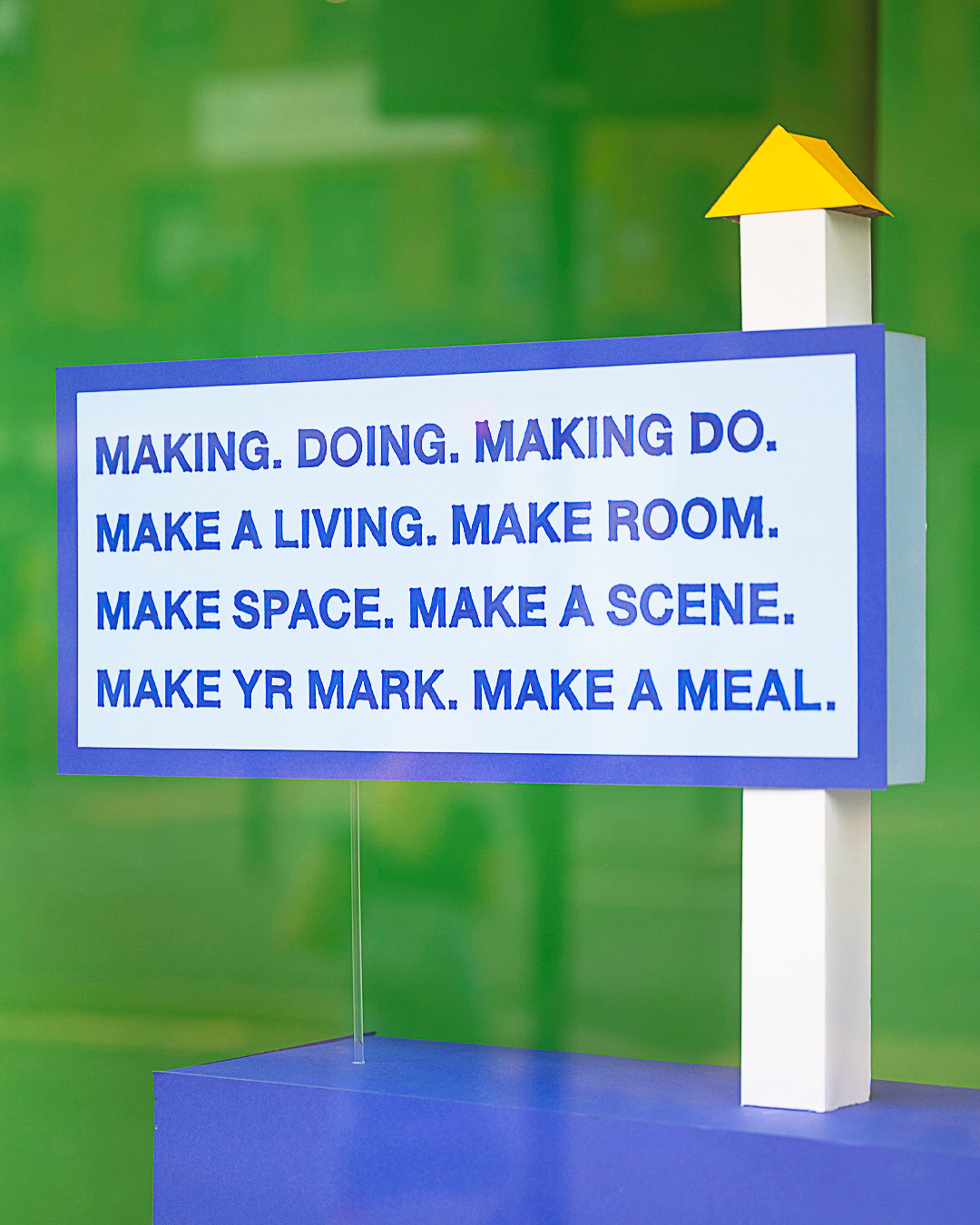 A blue and white sign with a yellow roof reads: MAKING. DOING. MAKING DO. MAKE A LIVING. MAKE ROOM. MAKE SPACE. MAKE A SCENE. MAKE YOUR MARK. MAKE A MEAL. The background is green.