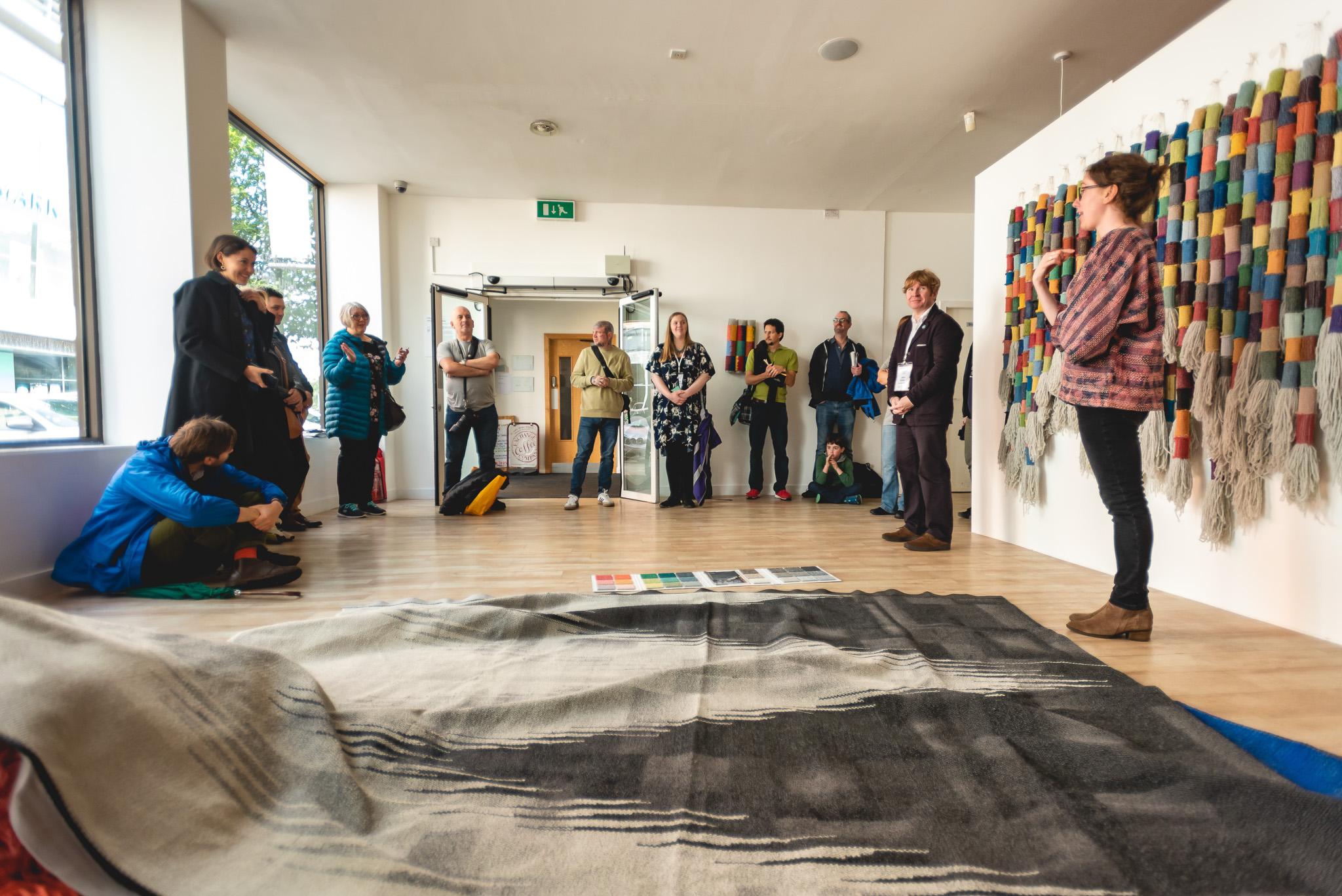 A group of people stand and sit in a bright gallery space, listening to a speaker near a colourful woven wall hanging. Textile artworks and samples are displayed on the wooden floor.