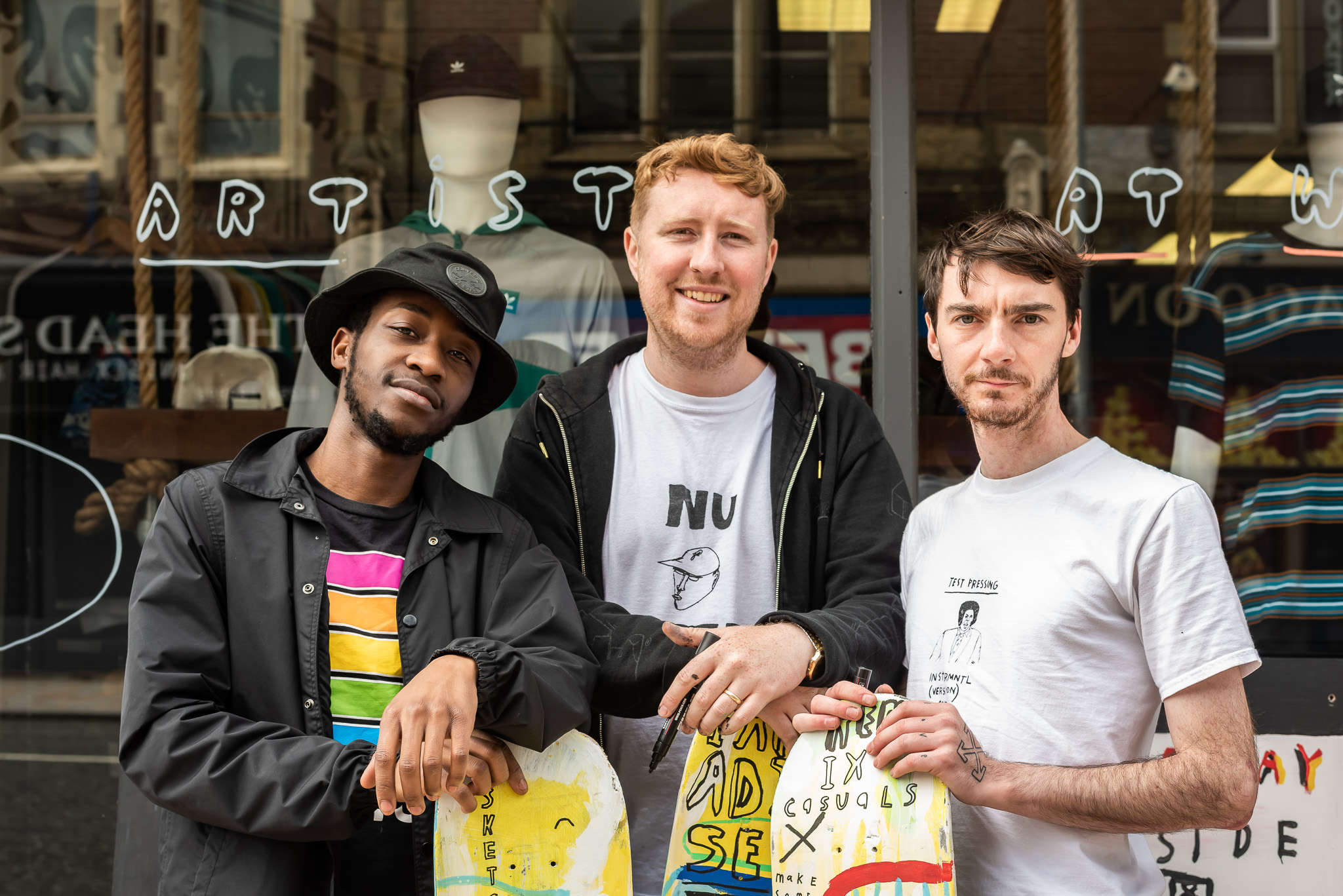 Three men stand together outside a shopfront with “ARTIST” on the window. They are smiling and holding printed artwork. One wears a hat and a striped shirt, the others wear white shirts.