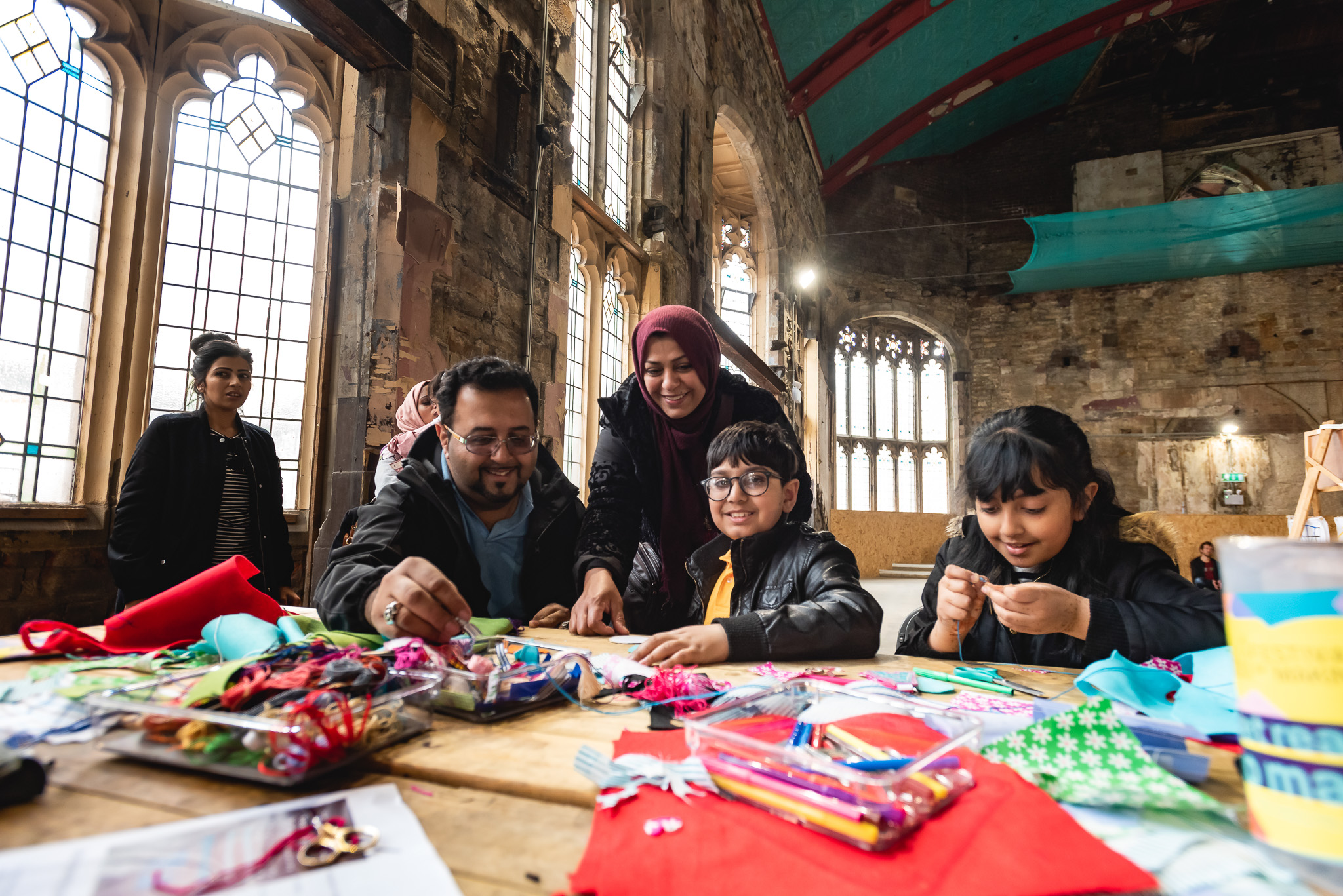 A family sits round a table in a large, sunlit room with stained glass windows, smiling and working together on colourful arts and crafts projects. Supplies and decorations cover the table.