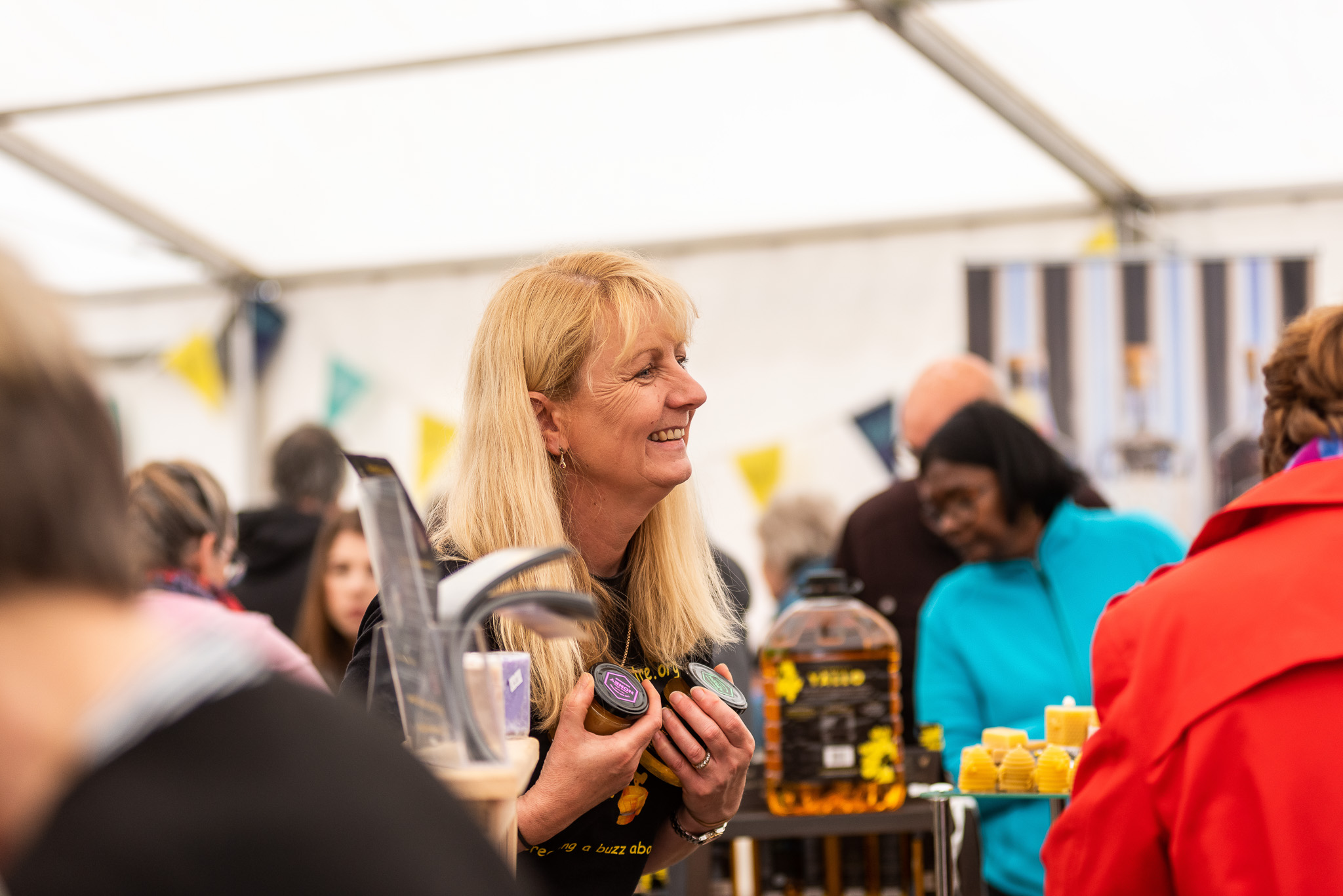 A smiling woman holds jars at a busy market stall under a marquee, surrounded by people shopping. Colourful bunting decorates the background, and products like honey and candles are displayed on the table.