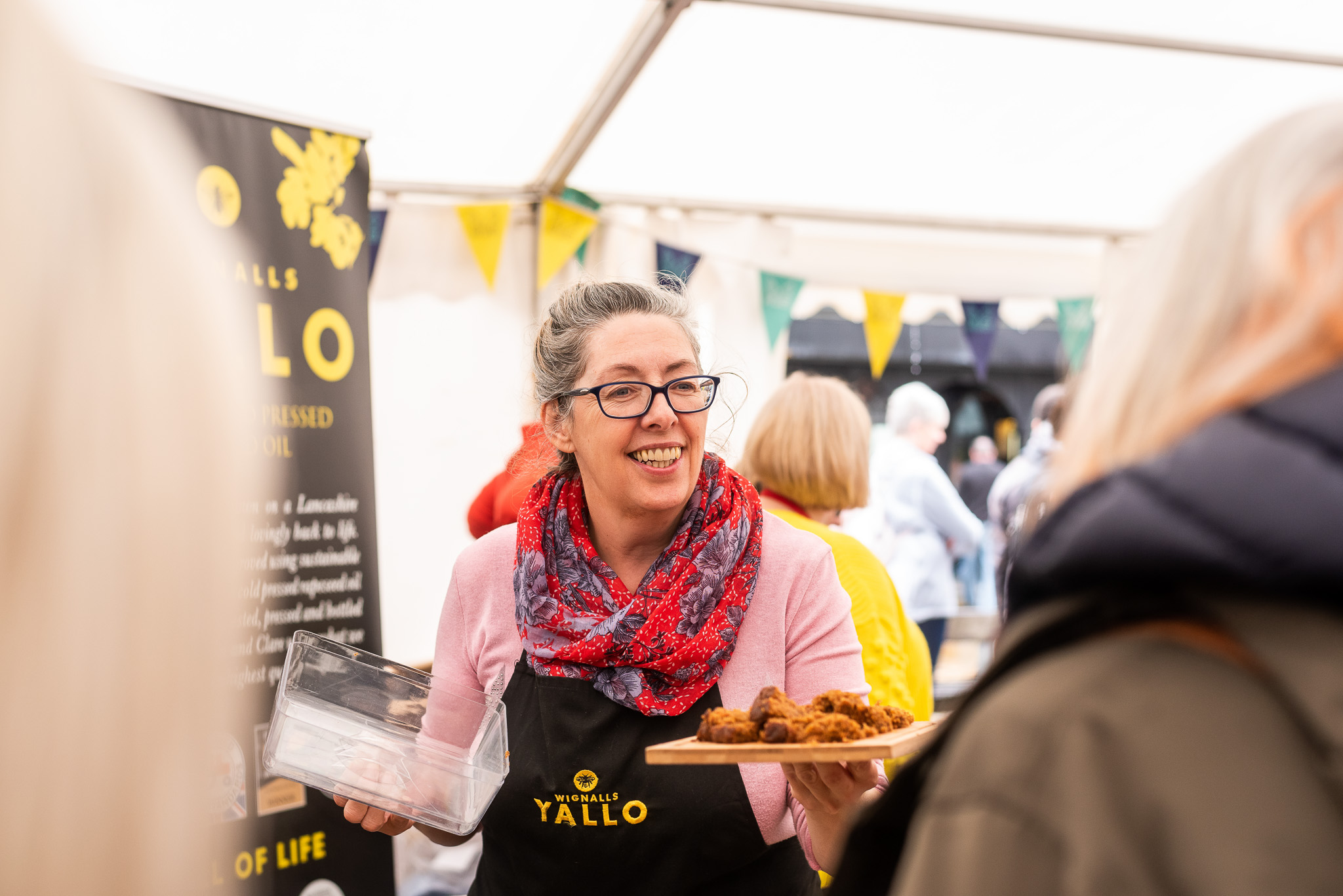 A smiling woman wearing glasses and a pink scarf offers food samples on a tray at a market stall. Colourful bunting decorates the tent, and people are gathered around her.