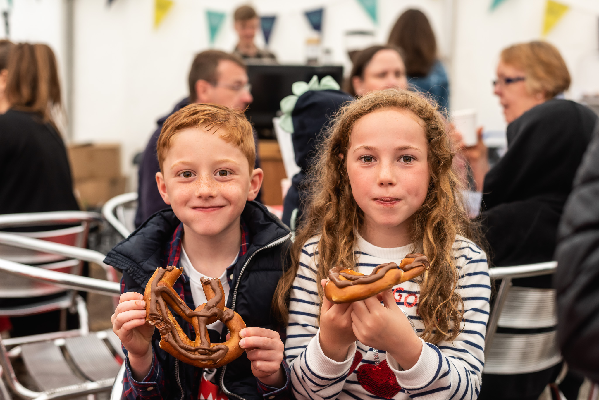 Two children, a boy and a girl, sit side by side indoors, smiling and holding large pretzels. People are sitting and talking in the blurred background. The scene appears lively and festive.