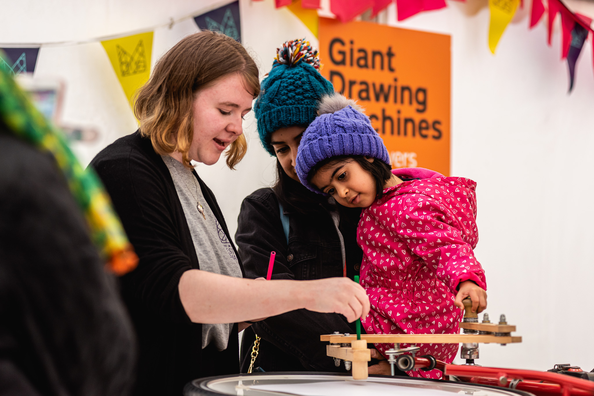 A woman helps a young child in a pink coat use a drawing machine, whilst another child in a blue hat watches. Colourful bunting and a sign reading Giant Drawing Machines are in the background.