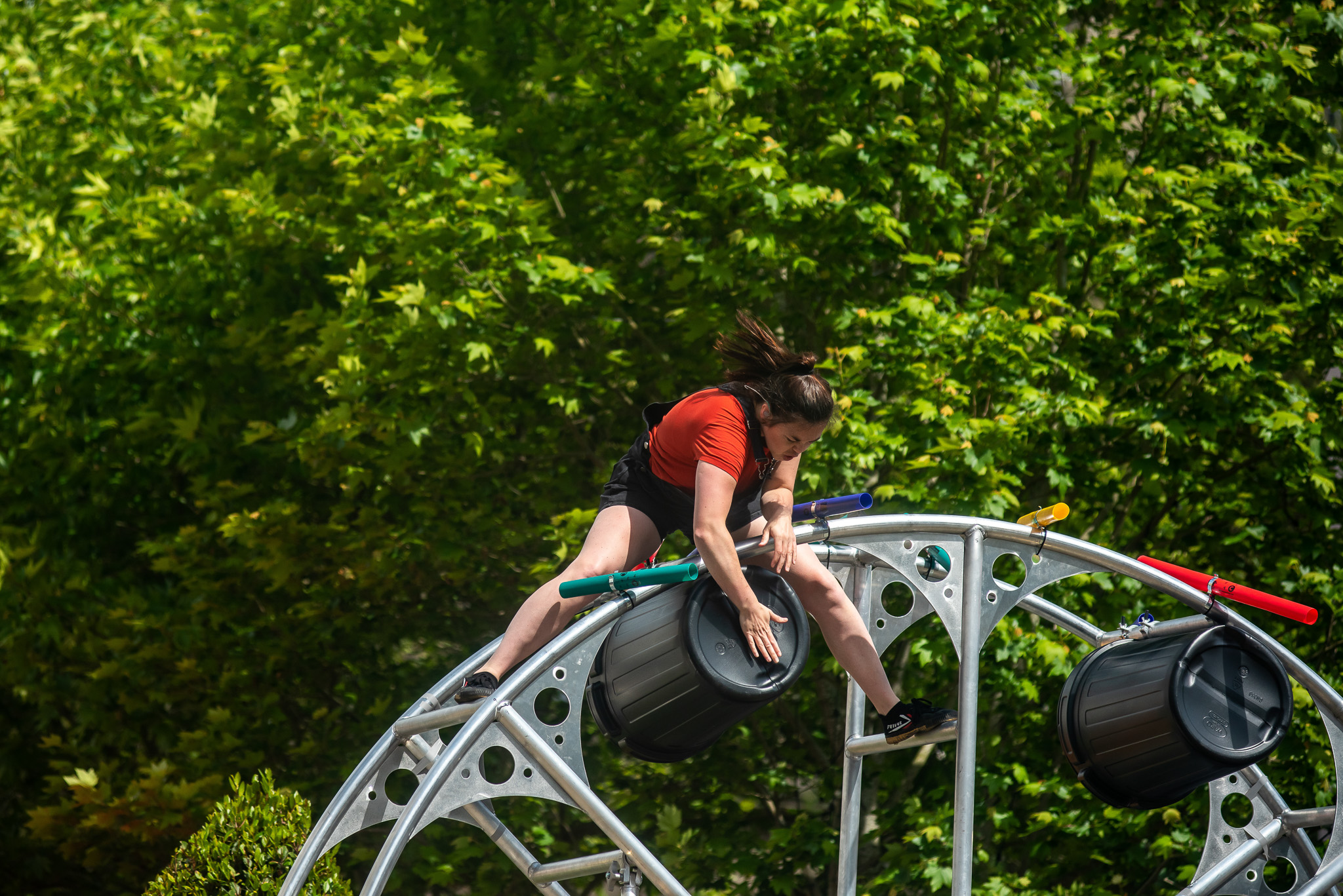 A person in a red shirt and black shorts climbs a metal structure holding a black drum, set against a background of lush green trees under sunlight.
