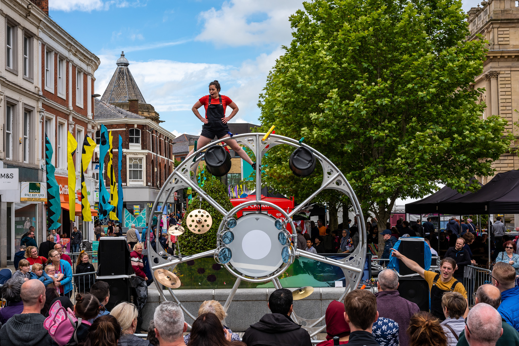 A performer stands on top of a large metal wheel structure during a lively outdoor street festival, surrounded by a crowd of spectators, colourful flags, and trees under a blue sky.