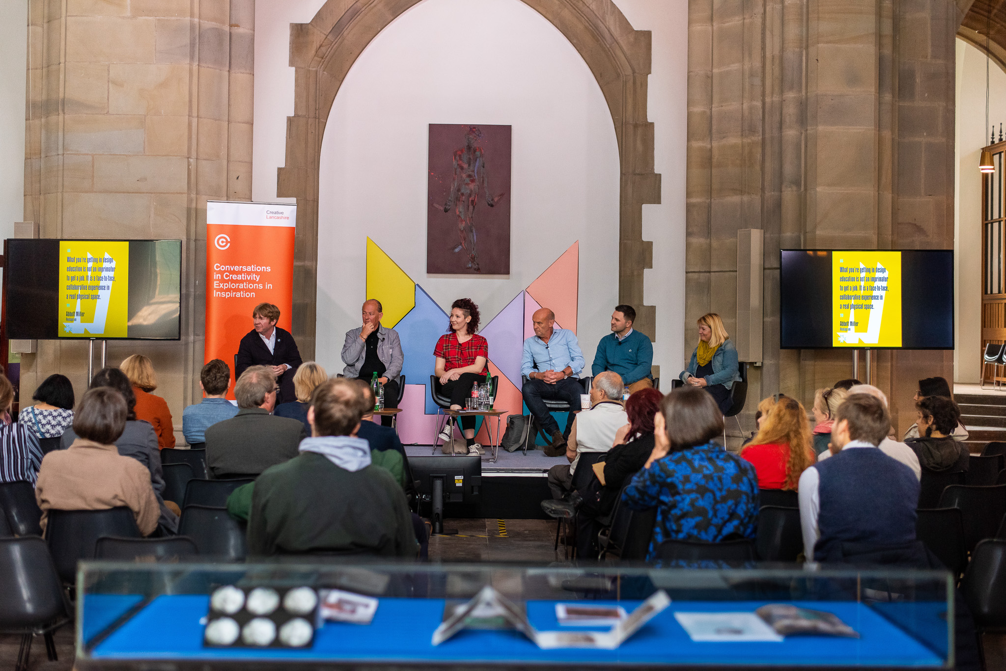 A panel of six people sits on a stage in front of an audience in a large room with arched stone architecture. Two screens display yellow graphics, and a banner reads “Conversations about collections, education, exploration, inspiration”.
