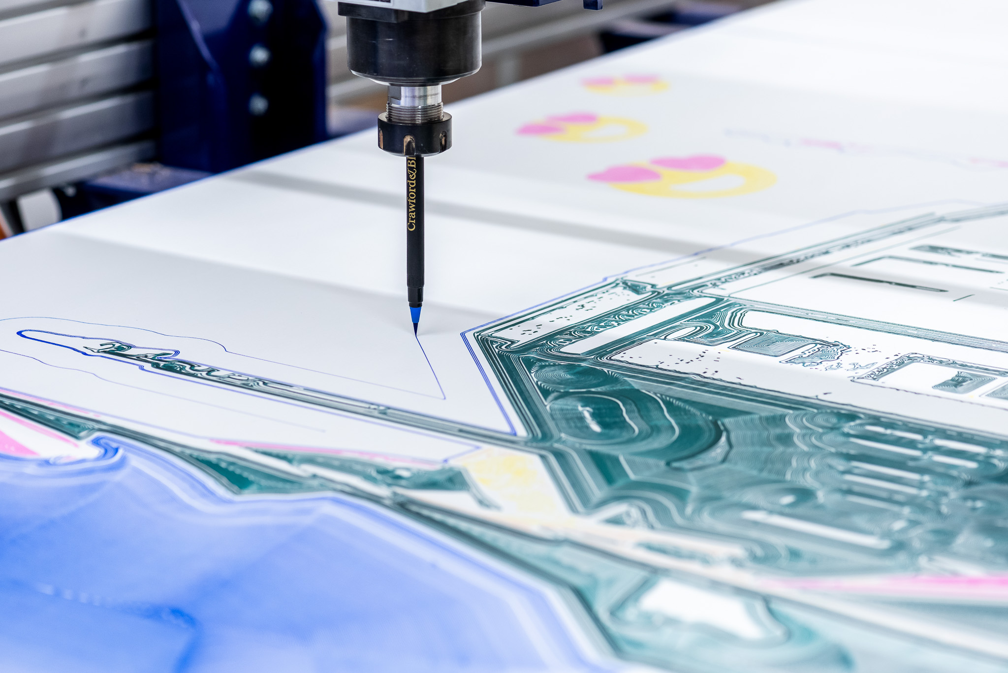 A close-up of a CNC machine using a blue-marking tool to outline shapes on a colourful, technical drawing or printed sheet, with mechanical details visible in the background.