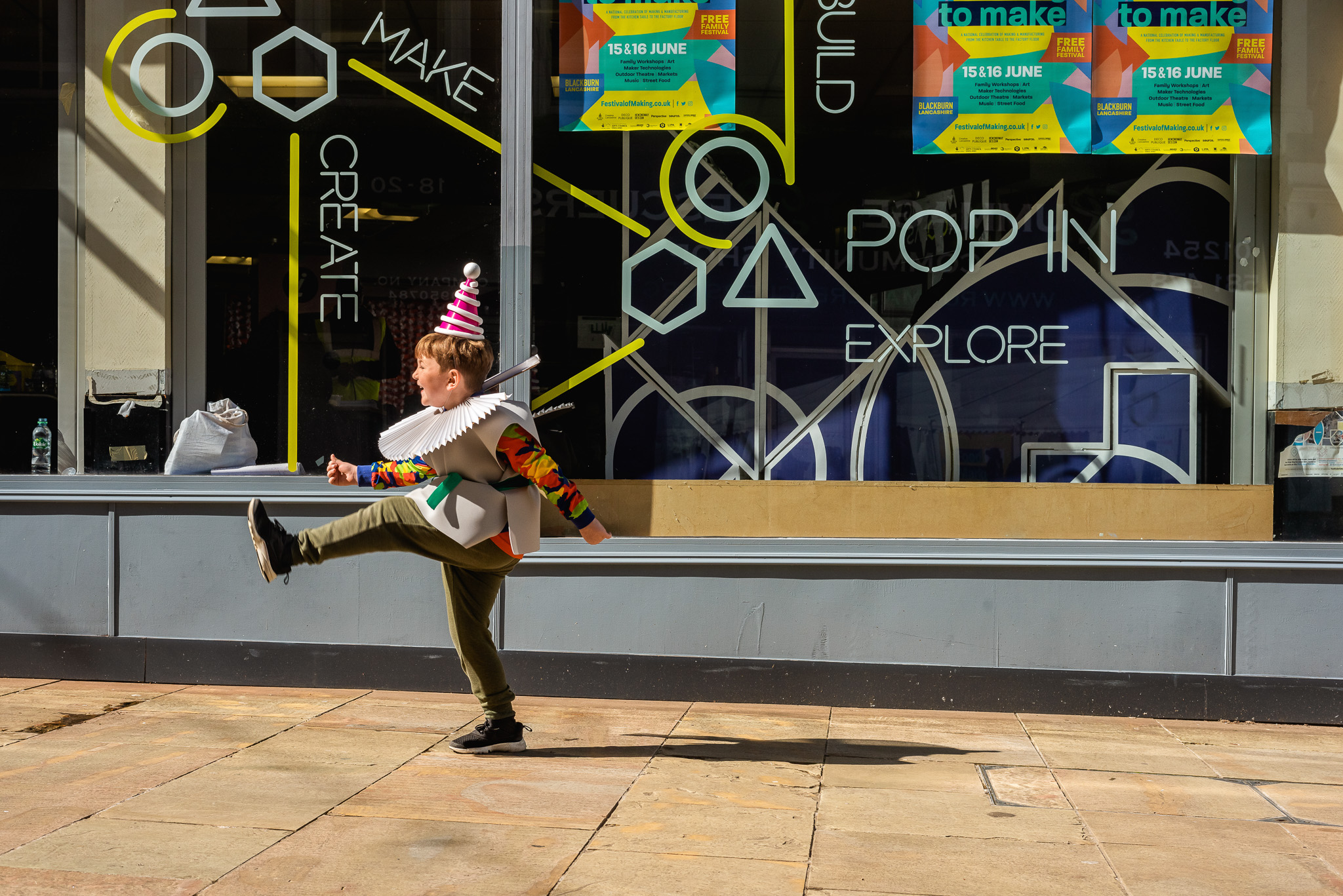 A child dressed in a colourful clown costume and party hat joyfully kicks one leg up whilst walking in front of a shop window decorated with geometric shapes and event posters.