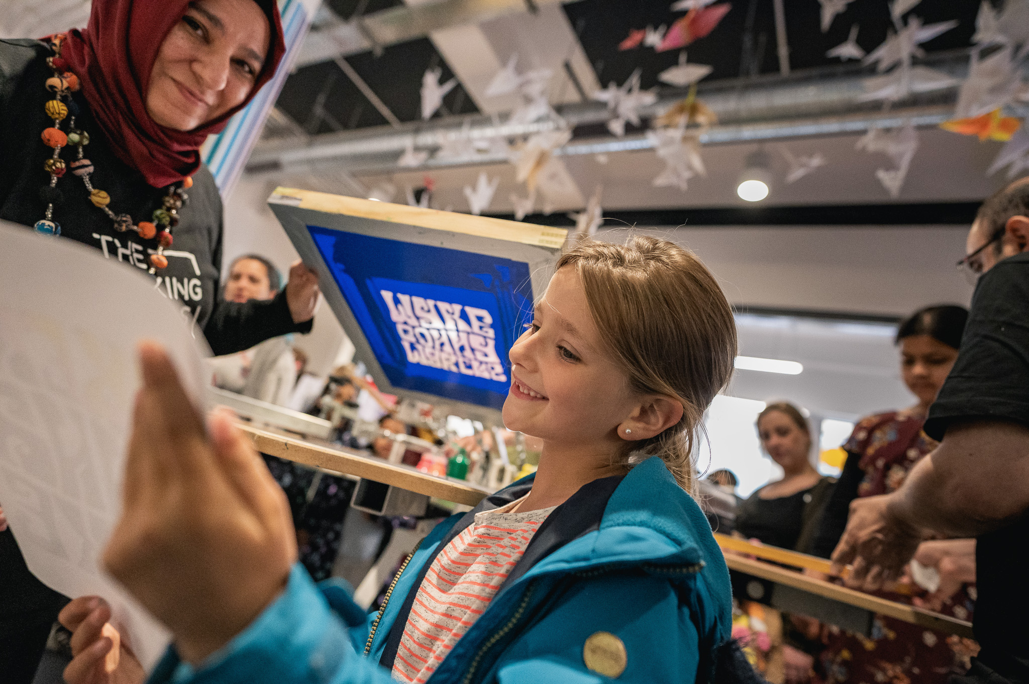 A smiling girl in a blue jacket examines a piece of paper as a woman holds a screen-printing frame nearby; other people and hanging paper cranes are visible in the background.