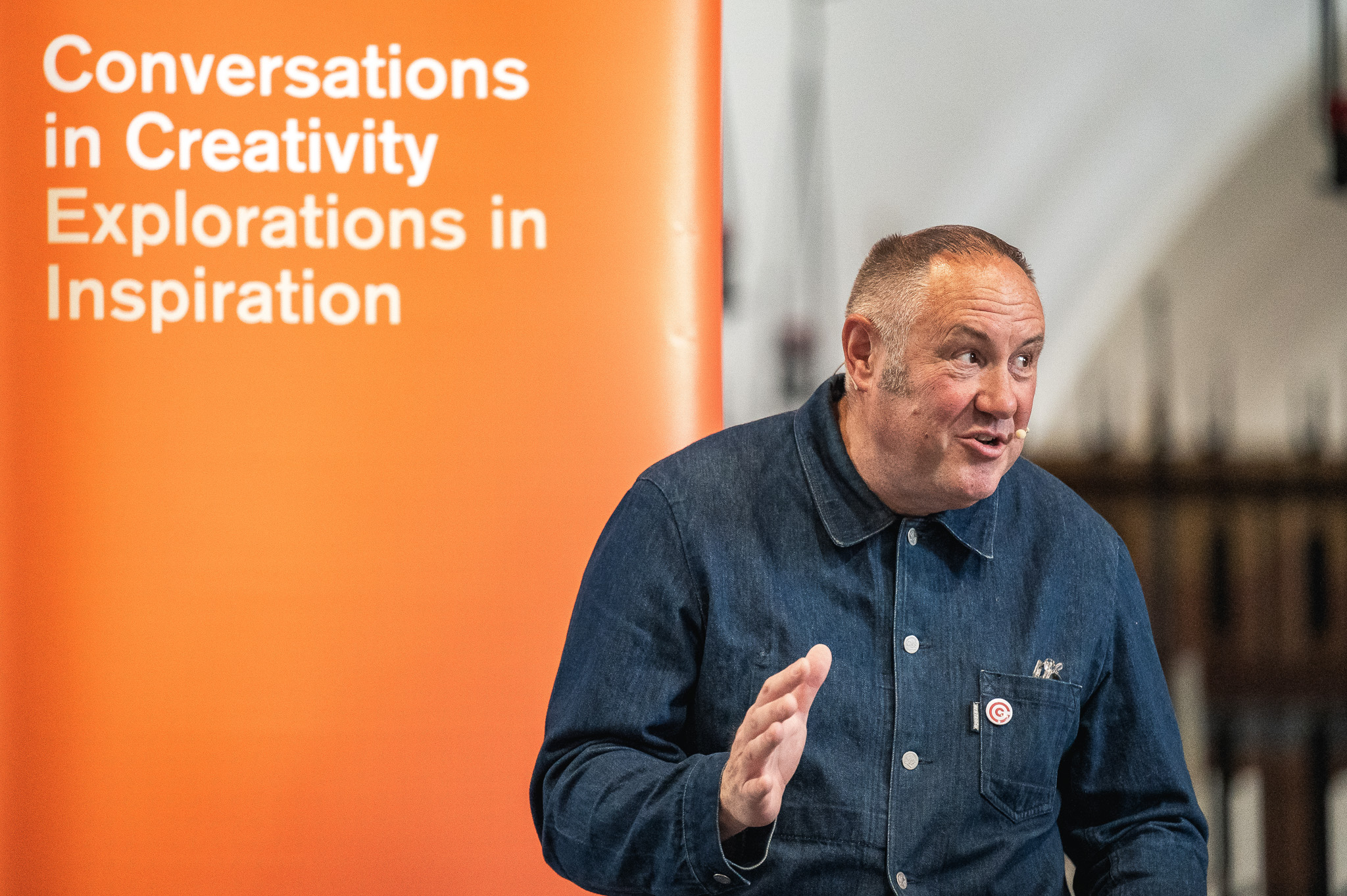 Keith Brymer Jones in a denim jacket gestures whilst speaking in front of an orange banner that reads, Conversations in Creativity, Explorations in Inspiration.