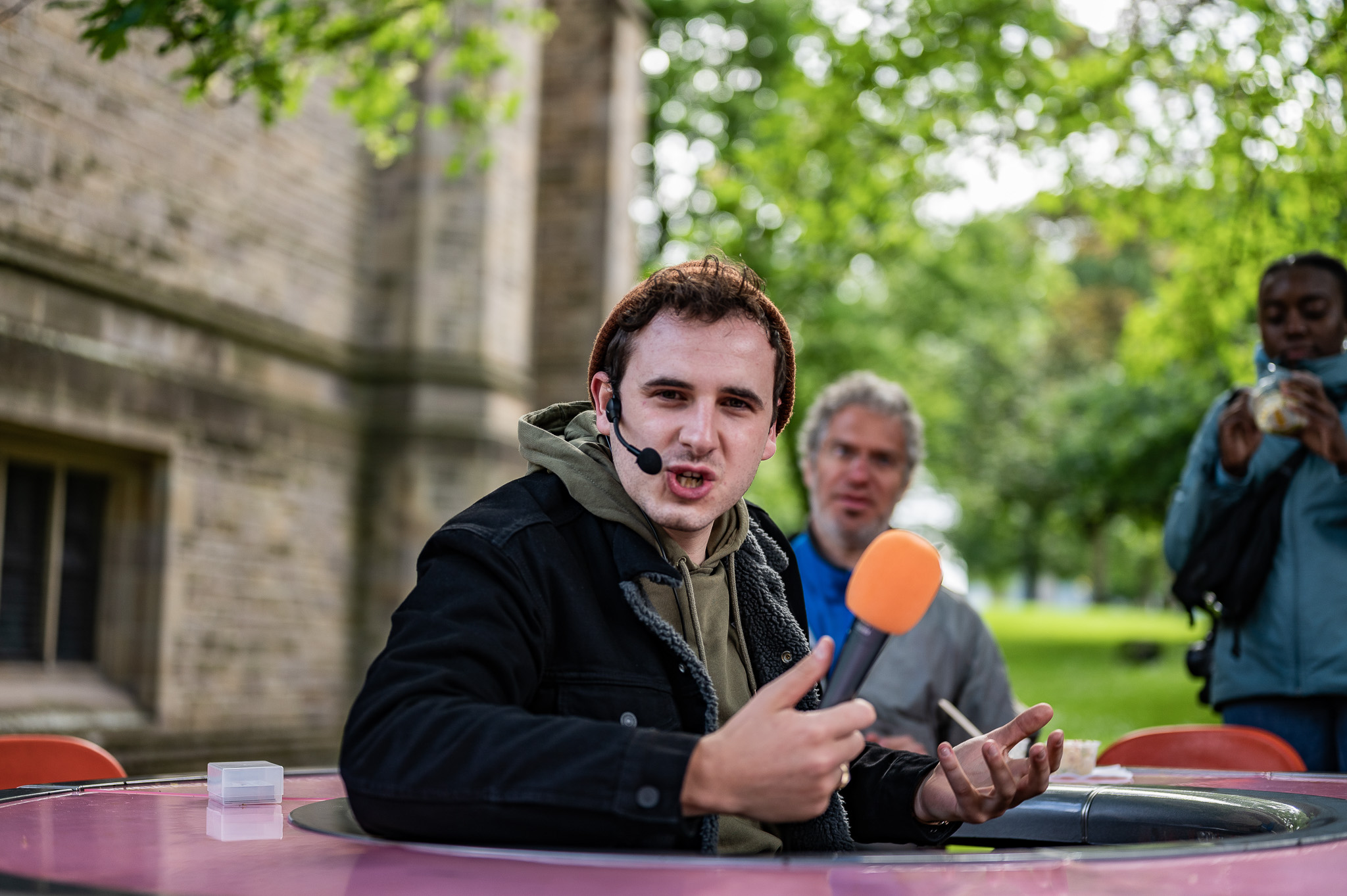 A young man wearing a headset and holding an orange microphone speaks animatedly whilst sitting at a round pink table outdoors, with two people and greenery in the background.