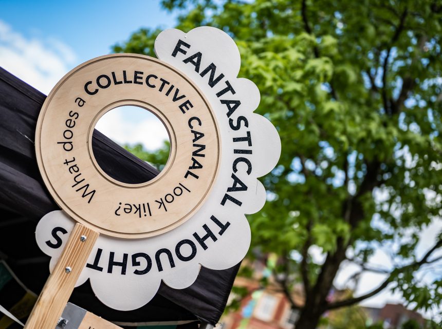 A round wooden sign with the words FANTASTICAL THOUGHTS and What does a COLLECTIVE CANAL look like? is displayed outdoors near trees and buildings.