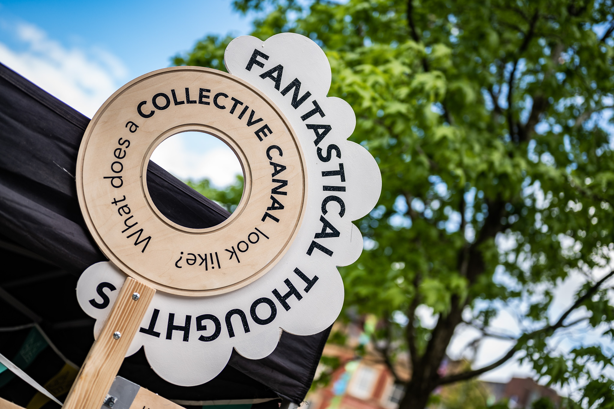 A round wooden sign with the words FANTASTICAL THOUGHTS and What does a COLLECTIVE CANAL look like? is displayed outdoors near trees and buildings.