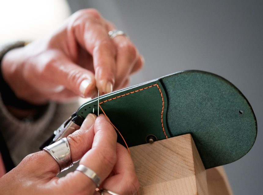 Close-up of hands with silver rings hand-stitching green leather with orange thread, using a needle and holding the leather piece steady on a wooden block.