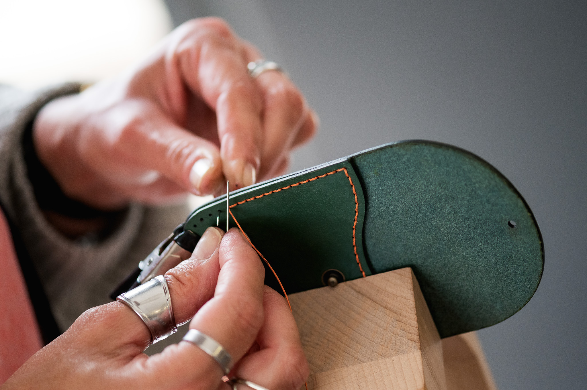 Close-up of hands with silver rings hand-stitching green leather with orange thread, using a needle and holding the leather piece steady on a wooden block.