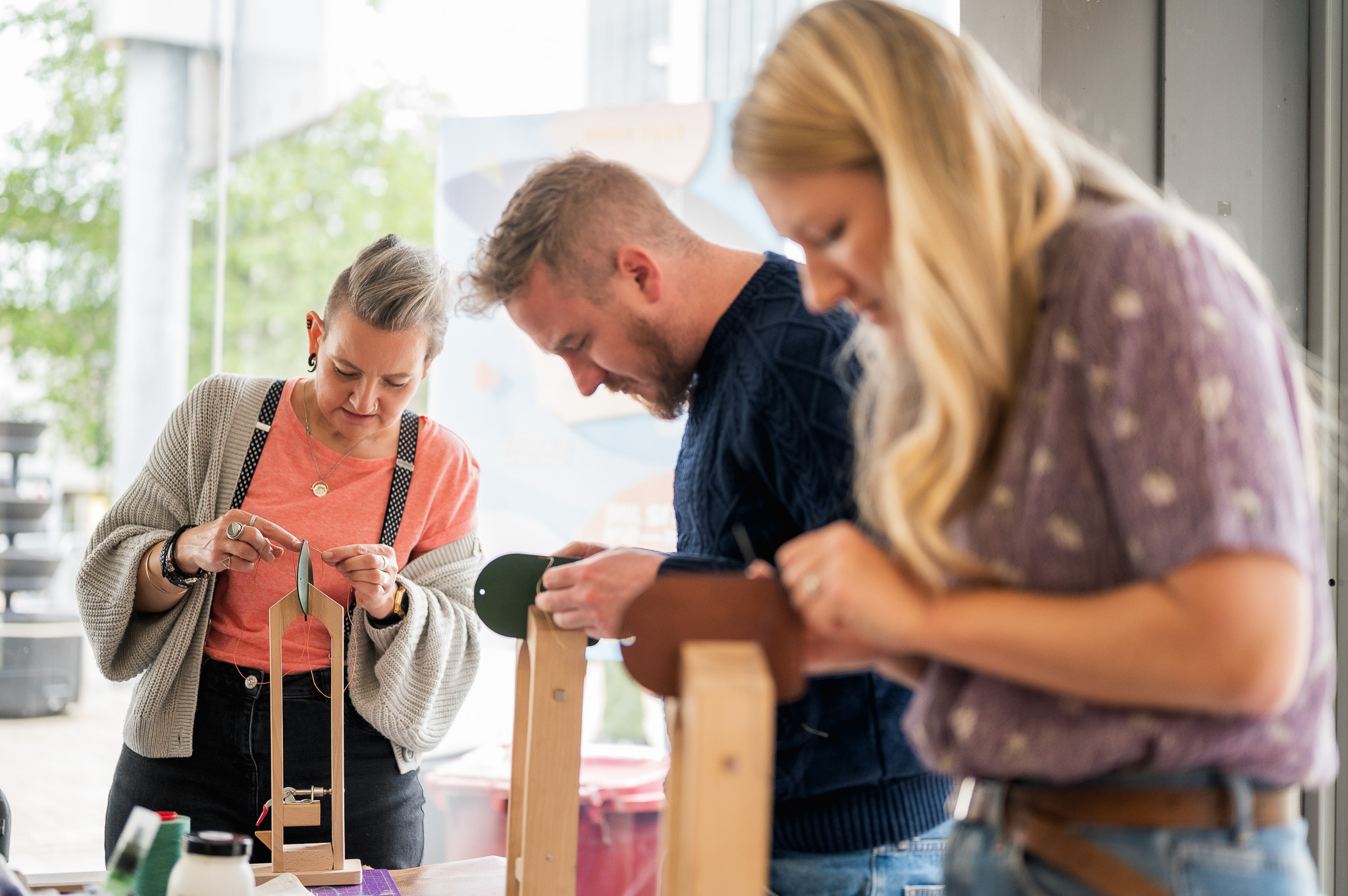 Three adults focus intently whilst working on crafts at a table indoors. They are using tools and wooden stands, appearing engaged in a hands-on creative activity.