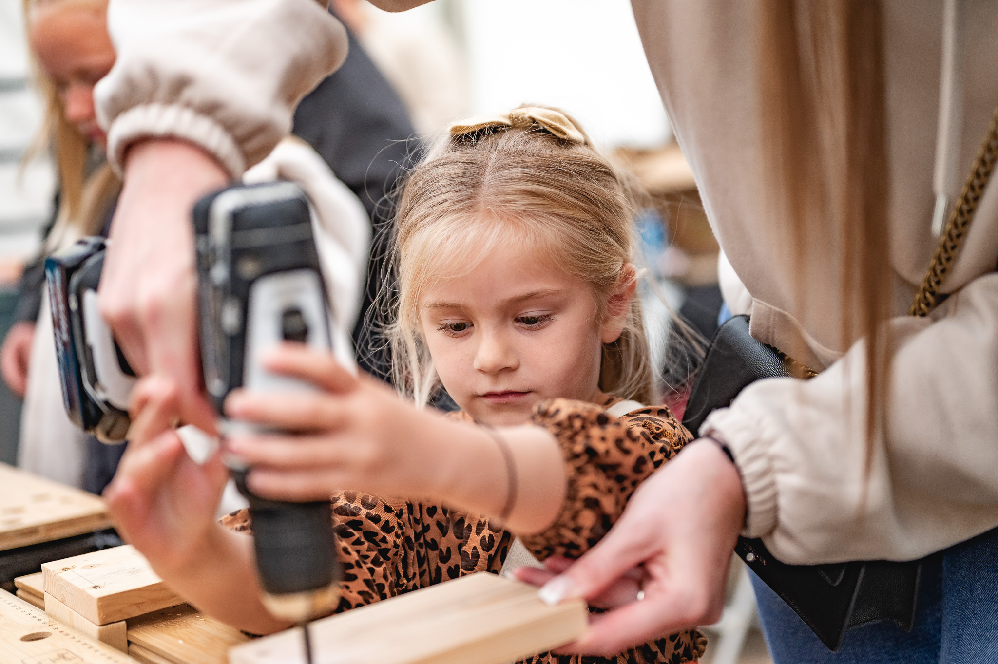 A young girl concentrating as an adult helps her use a power drill to work on a piece of wood. They are both focused on the task in a hands-on crafting or woodwork activity.