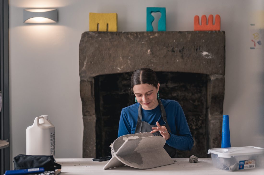 A woman wearing a blue shirt and apron works on a clay sculpture at a table, smiling. Behind her is a stone fireplace with colourful abstract decorations on the mantelpiece. Art supplies are on the table.
