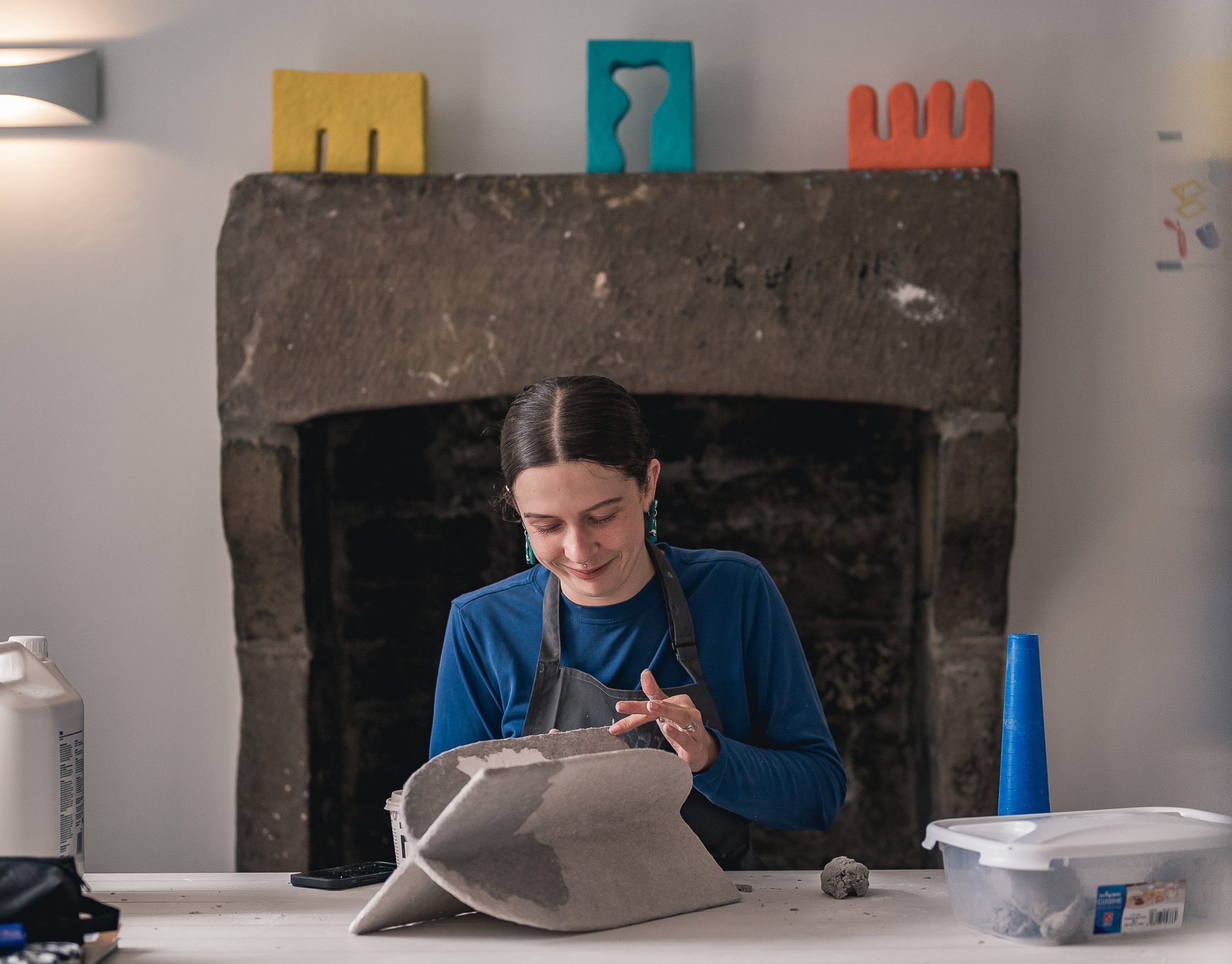 A person wearing a blue shirt and apron works on a sculptural piece at a table, with colourful abstract shapes displayed on a dark stone fireplace in the background. Art supplies are scattered on the table.
