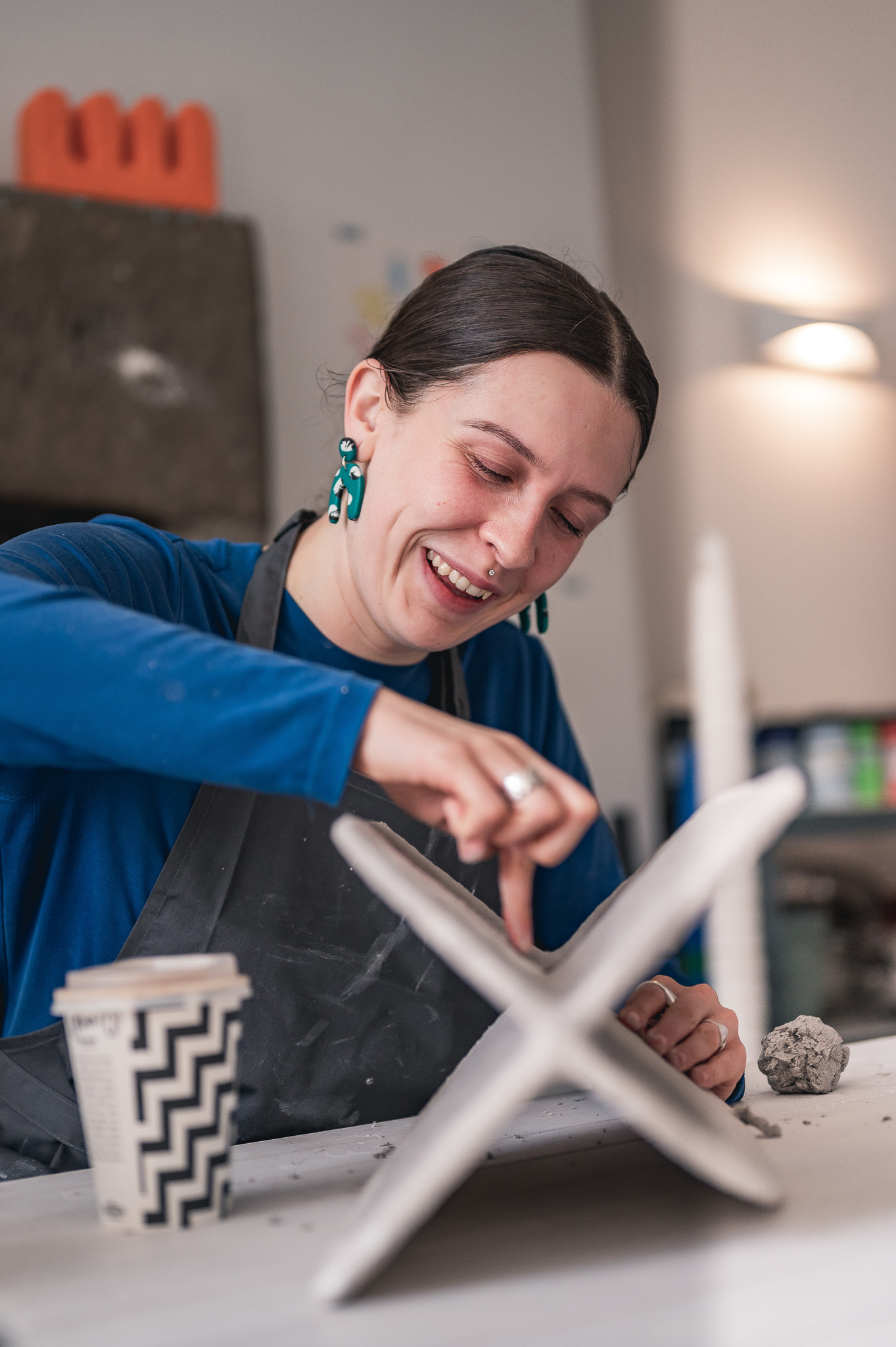 A woman wearing a blue shirt and apron smiles whilst sculpting a clay piece. She works at a table with a coffee cup and clay ball, in a well-lit art studio.