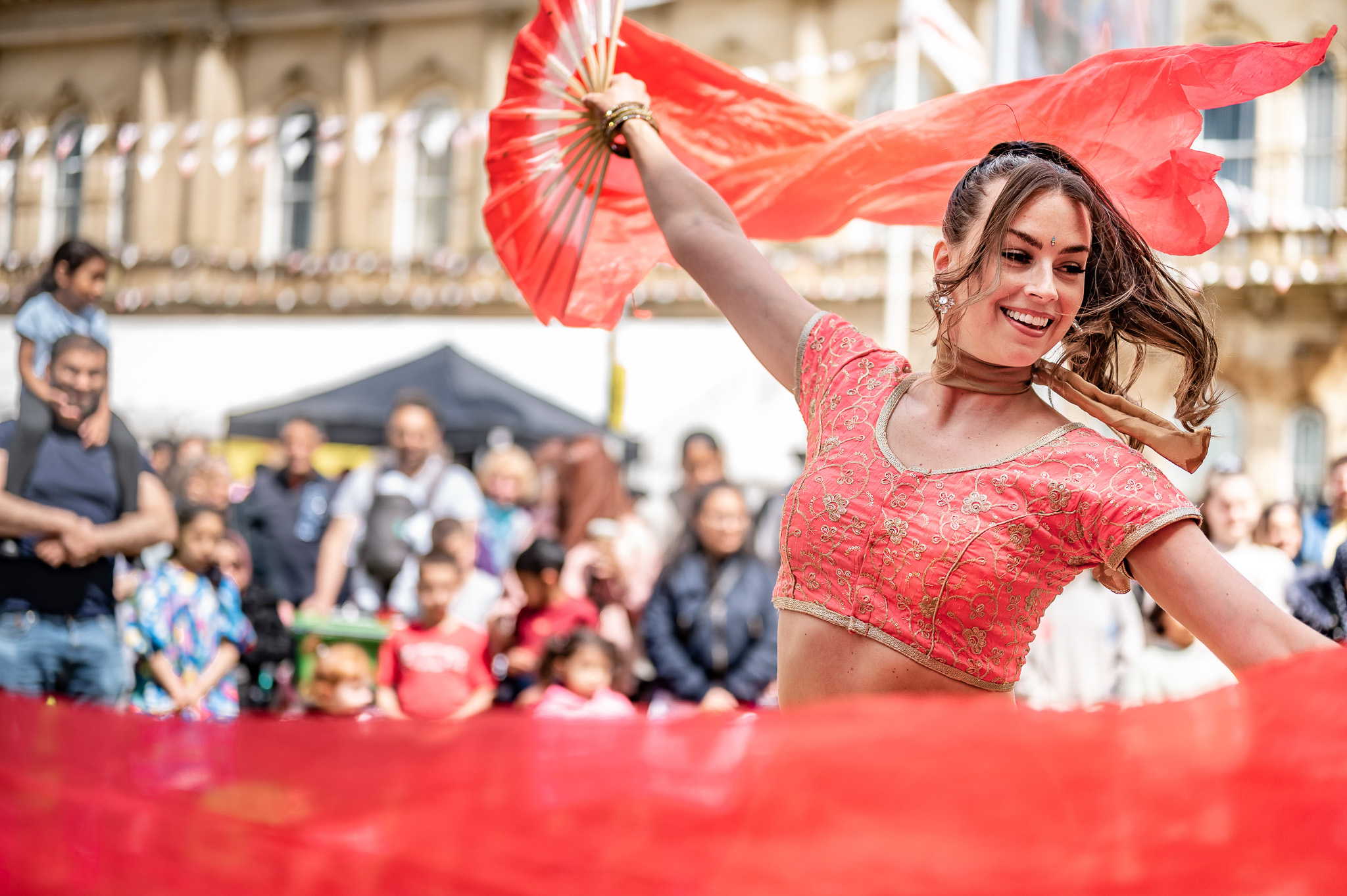 A woman in a vibrant red outfit dances joyfully with a red fan and scarf, smiling, as a crowd watches her outdoors. The background shows an historic building and onlookers enjoying the performance.