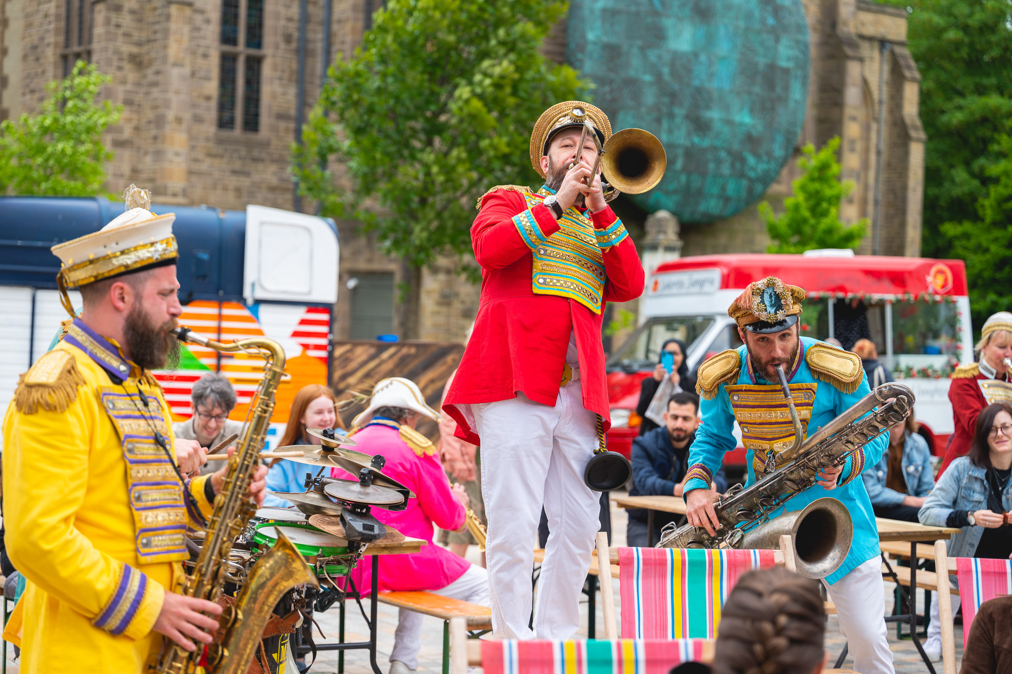 A lively outdoor performance featuring three musicians in colourful, ornate uniforms playing brass instruments, with a crowd of people watching. Trees, buildings, and food vans are visible in the background.