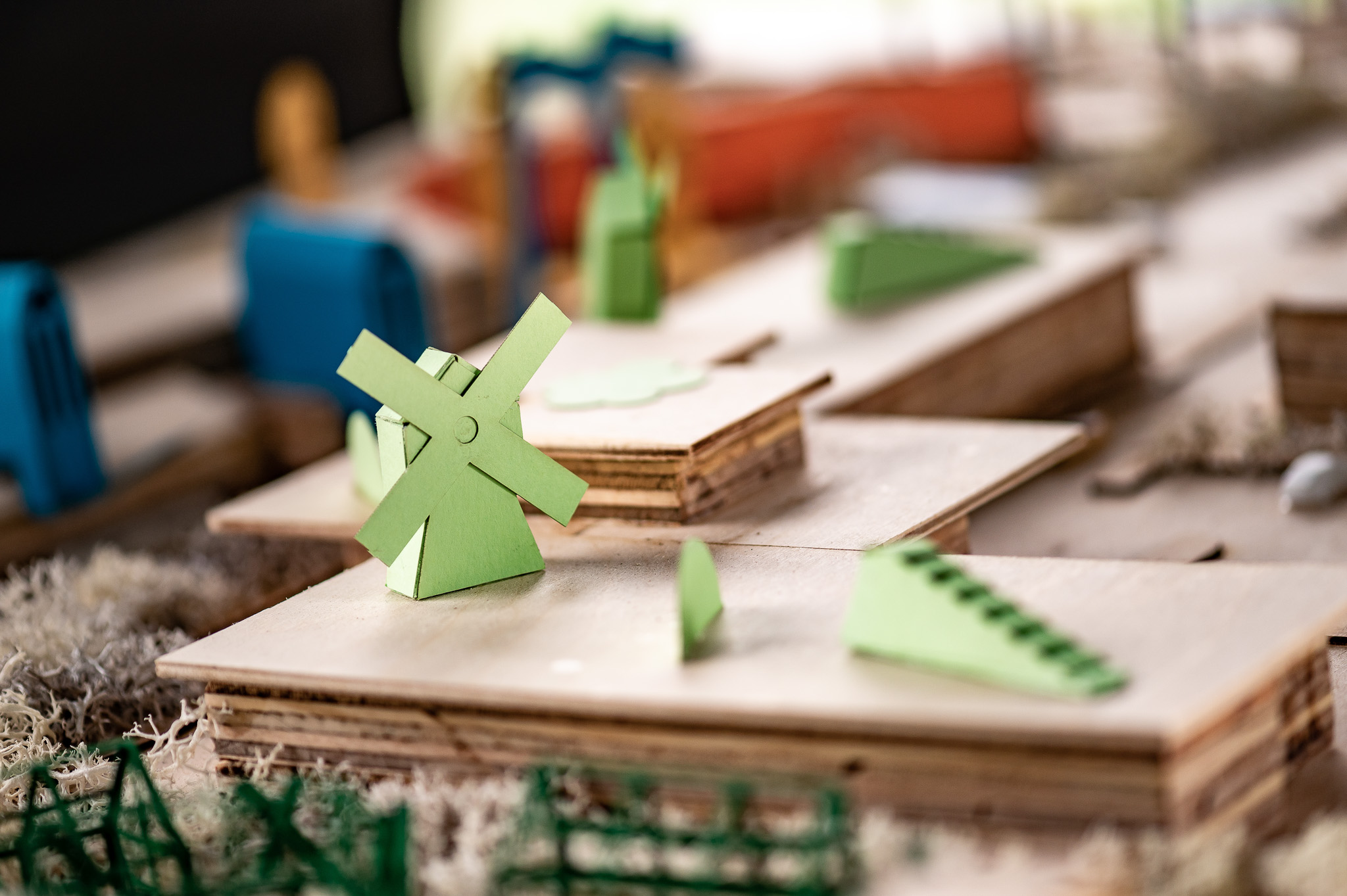 A close-up of a miniature architectural model featuring wooden platforms, a small green windmill, green triangular shapes, and various colourful abstract structures in the background.