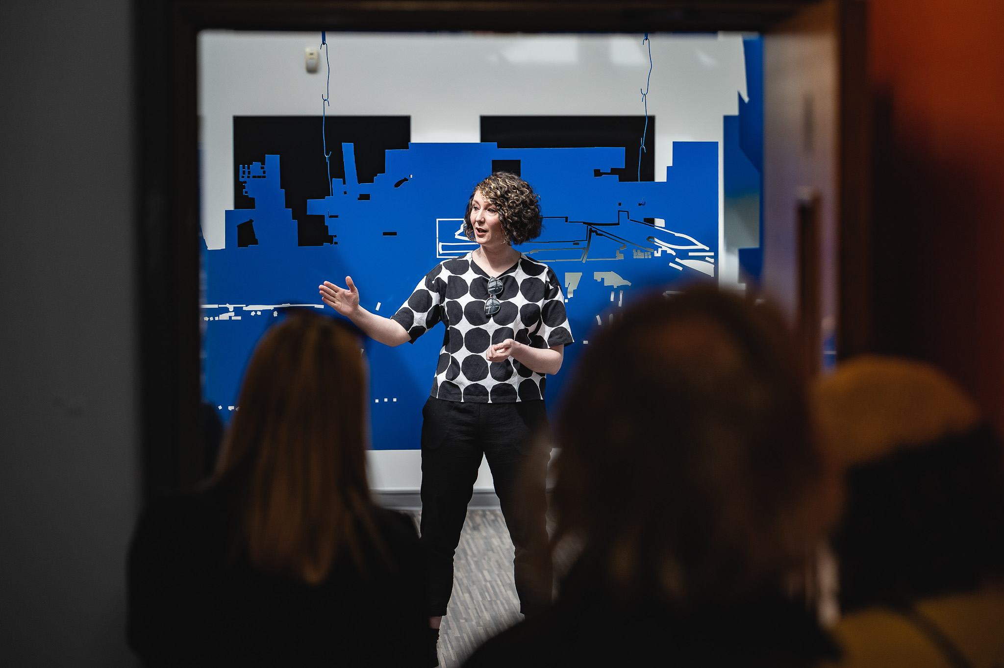 A person with curly hair, wearing a black and white spotty shirt, stands and gestures while speaking to an audience in a room with blue abstract wall art. The photo is taken from behind the seated audience.