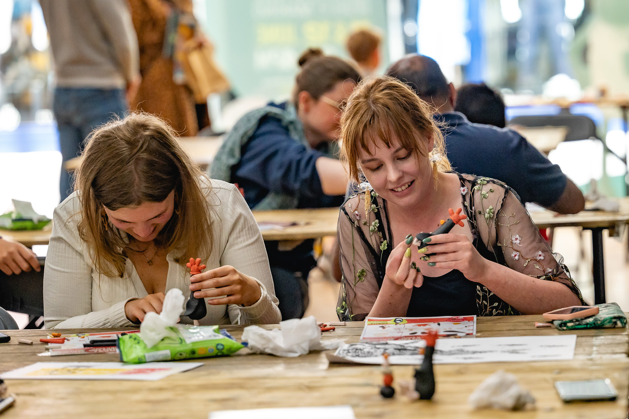 Two young women sit at a table, focused on crafting small figurines. Art supplies, wipes, and paper are spread out on the table. Other people in the background are also engaged in crafting activities.
