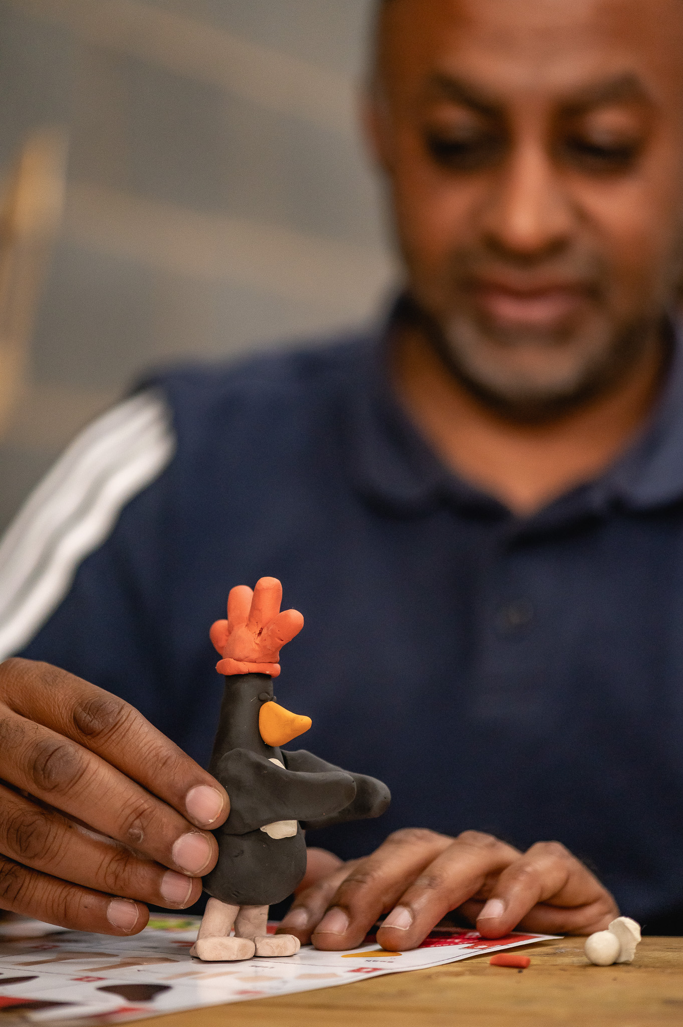 A person shapes a small penguin figurine with a red comb and yellow beak from modelling clay, focusing intently as their hands mould the details on a table.