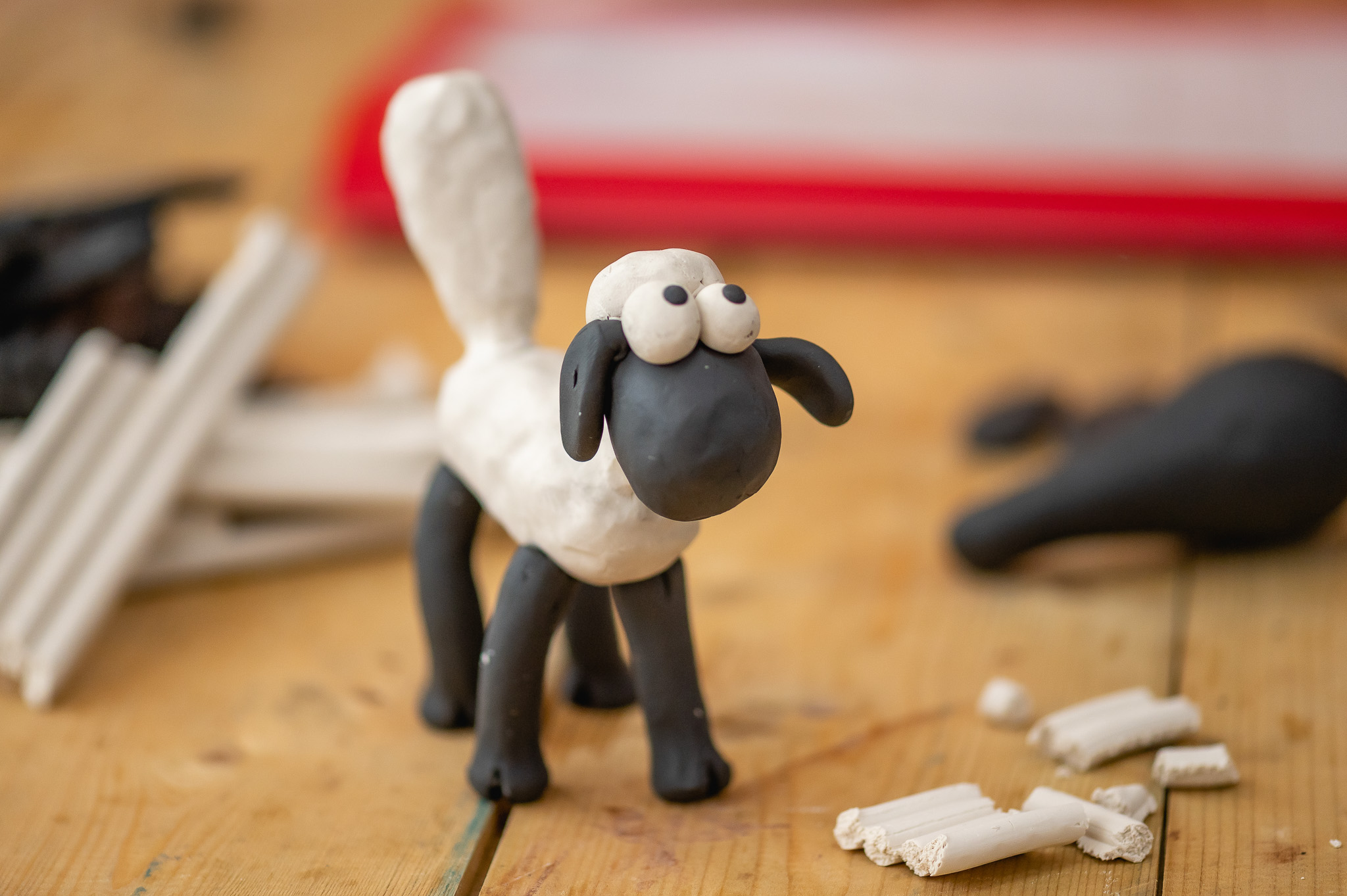 A small sheep figurine made of black and white clay stands on a wooden surface, with bits of white clay and other clay pieces scattered about.