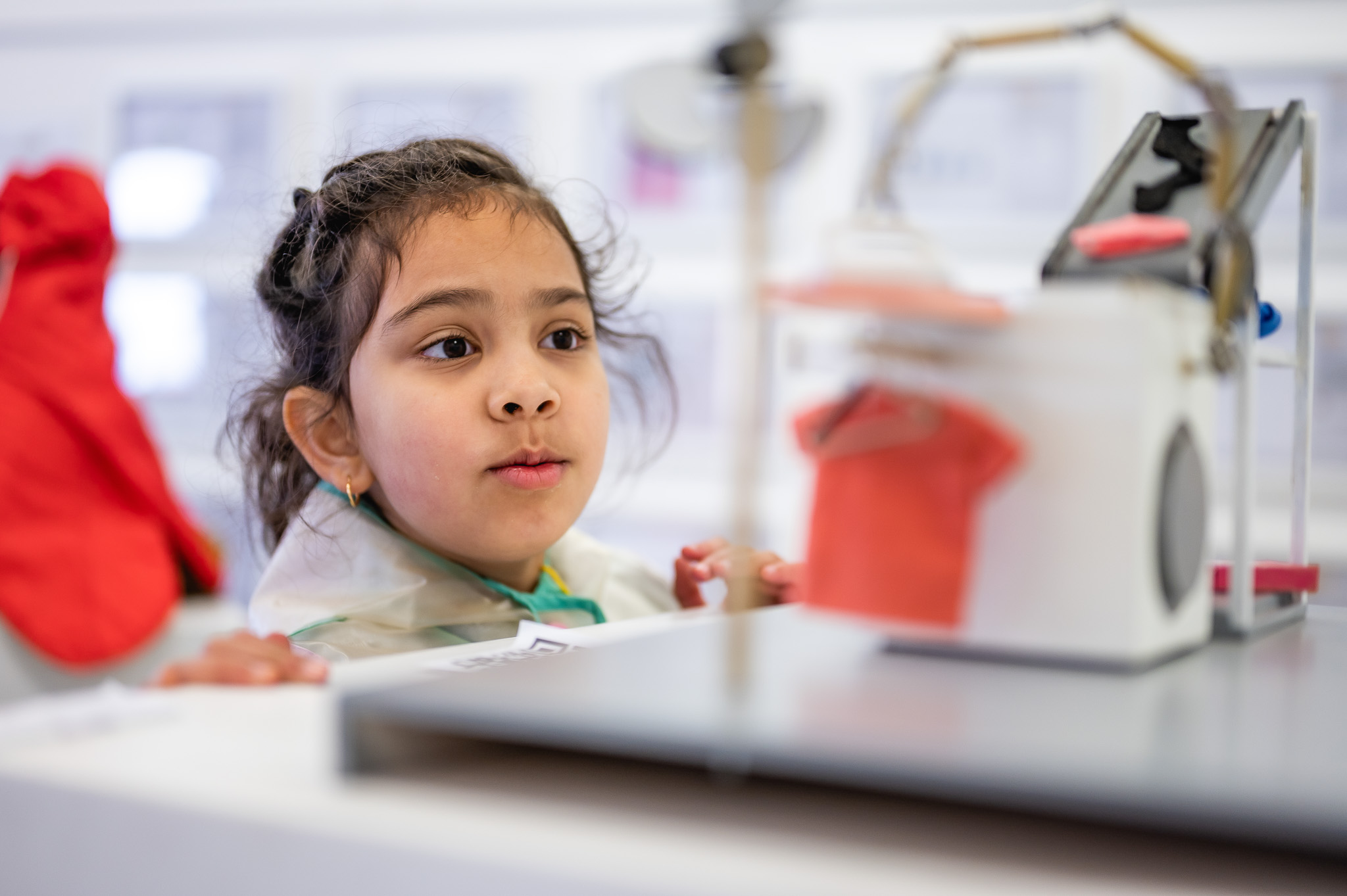 A young girl with dark hair and a curious expression observes a small mechanical model with miniature clothing and a washing machine in a brightly lit room.